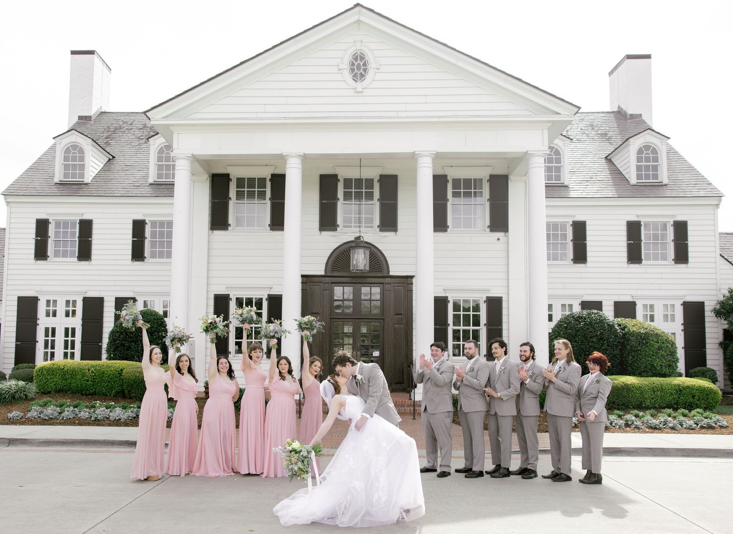 Wedding party poses in front of elegant white colonial mansion, bridesmaids in pink gowns and groomsmen in gray suits.