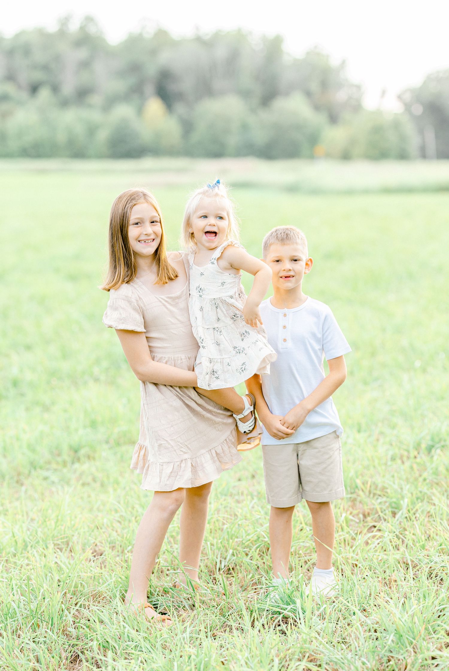 Three smiling children pose in a green field, with an older girl holding a toddler beside a young boy.