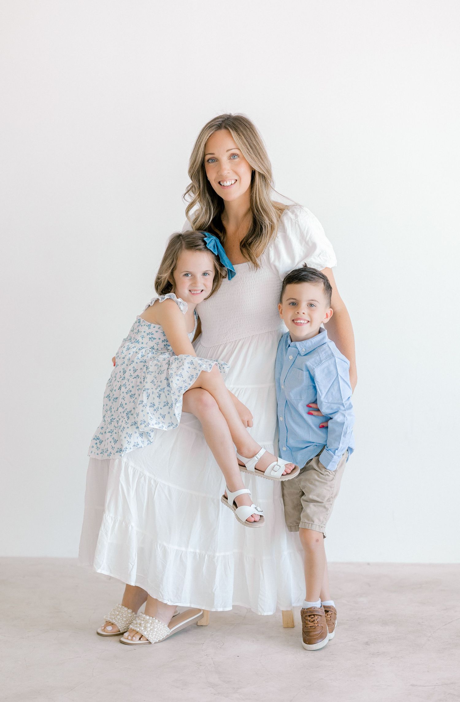 A smiling woman in a white dress poses with two young children in a bright, airy studio setting.