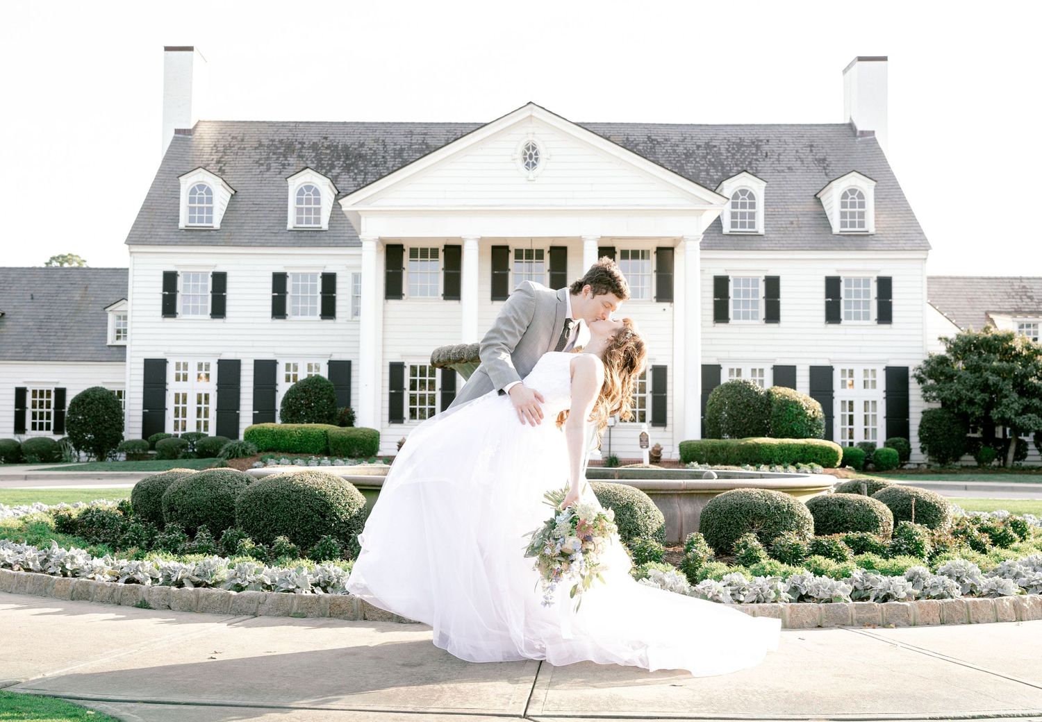 White colonial style mansion venue with landscaped gardens and long driveway on sunny wedding day.