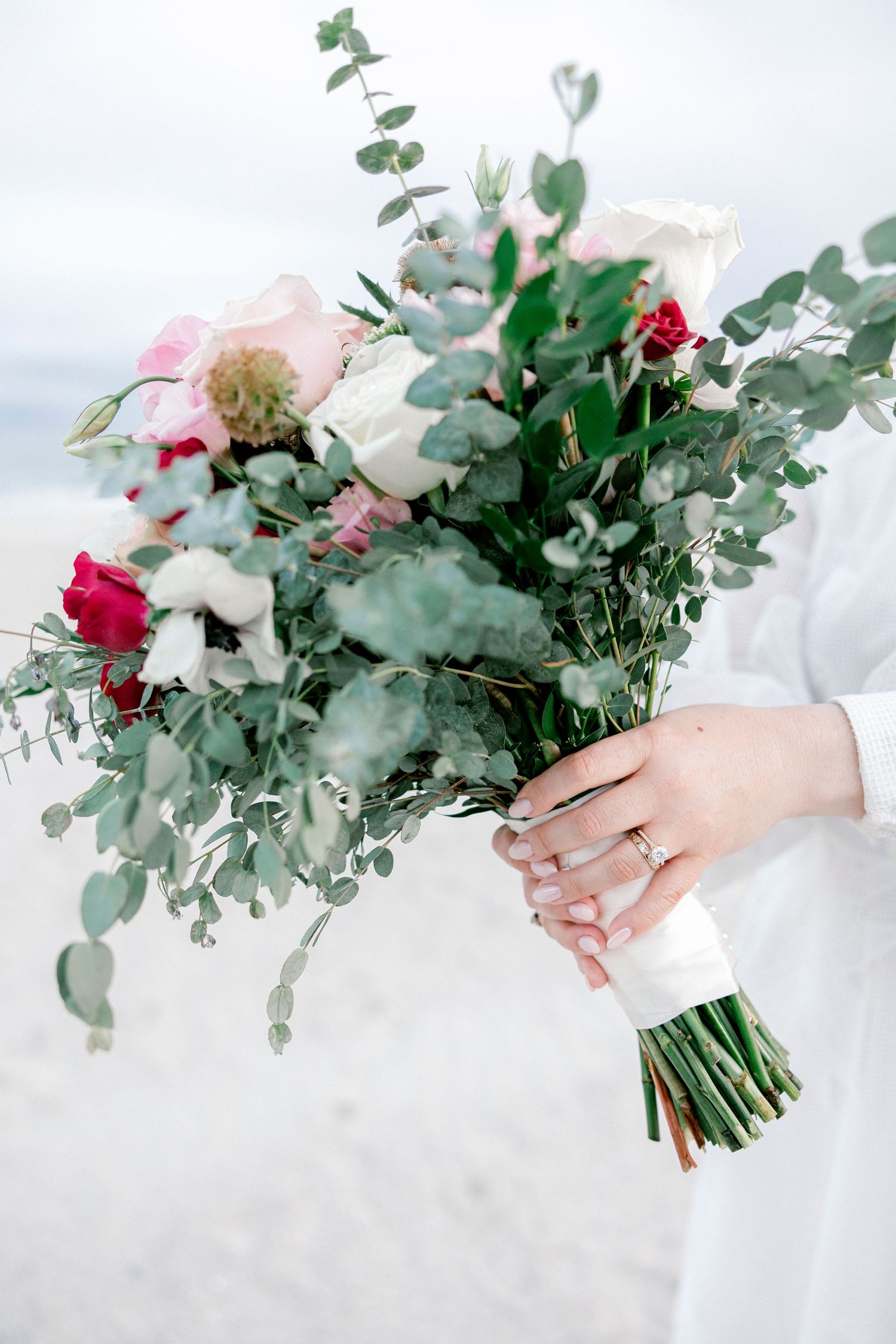 Bridal bouquet featuring pink and red roses with eucalyptus held against white dress.