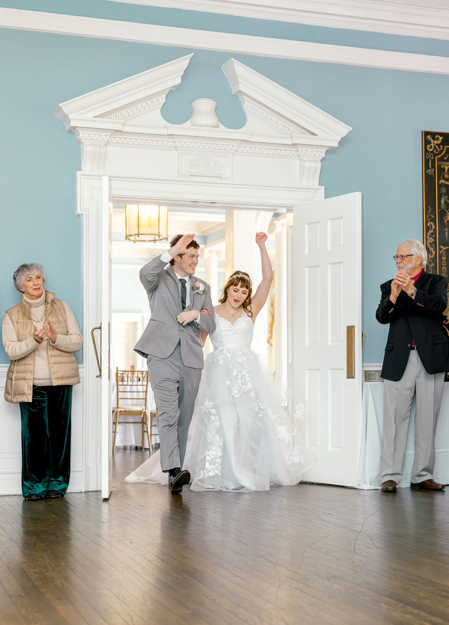 Wedding party members celebrate with energetic dance moves in a formal blue and white room.