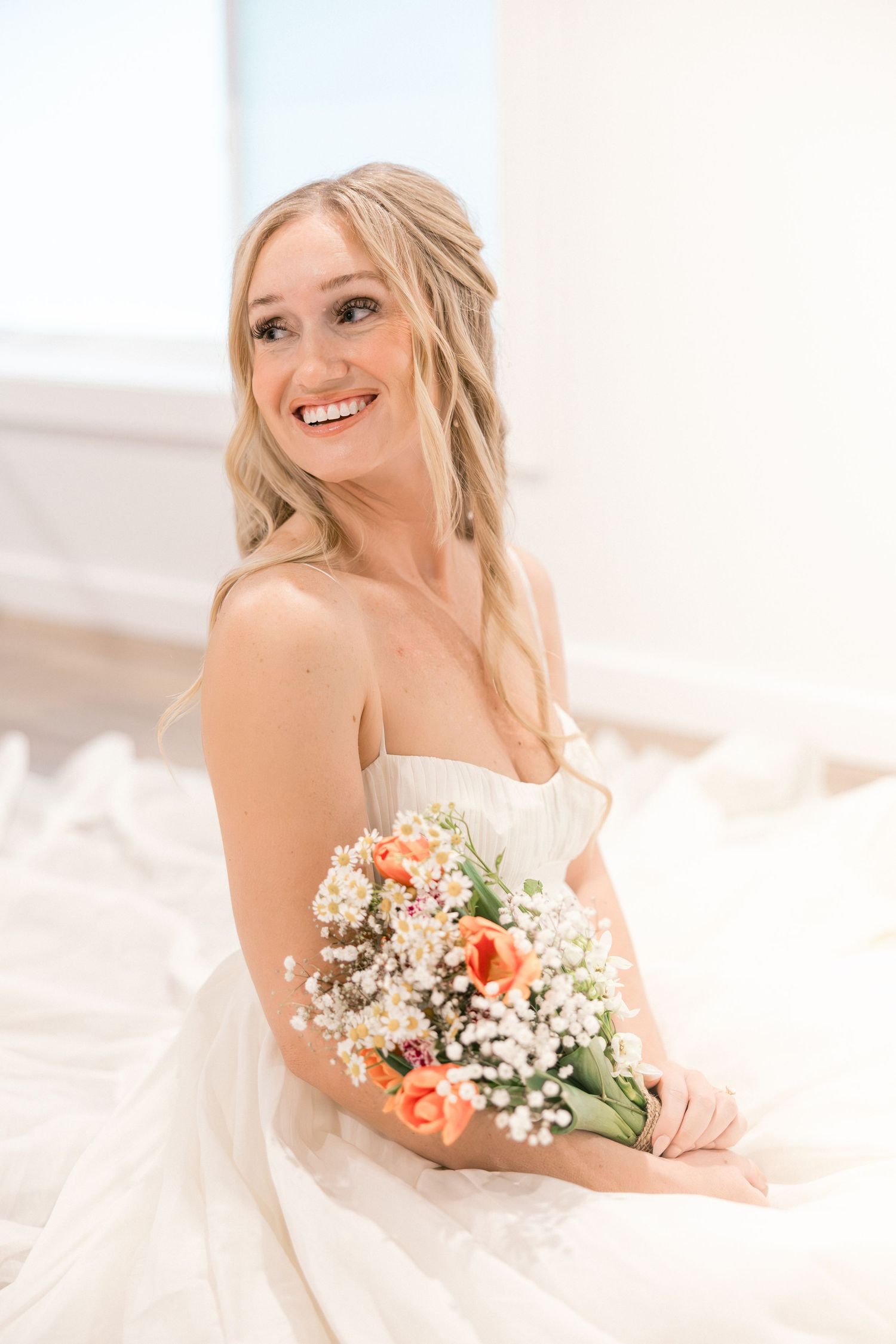 Smiling bride in strapless white gown holding a bouquet of peach tulips and baby's breath in a bright, airy setting.