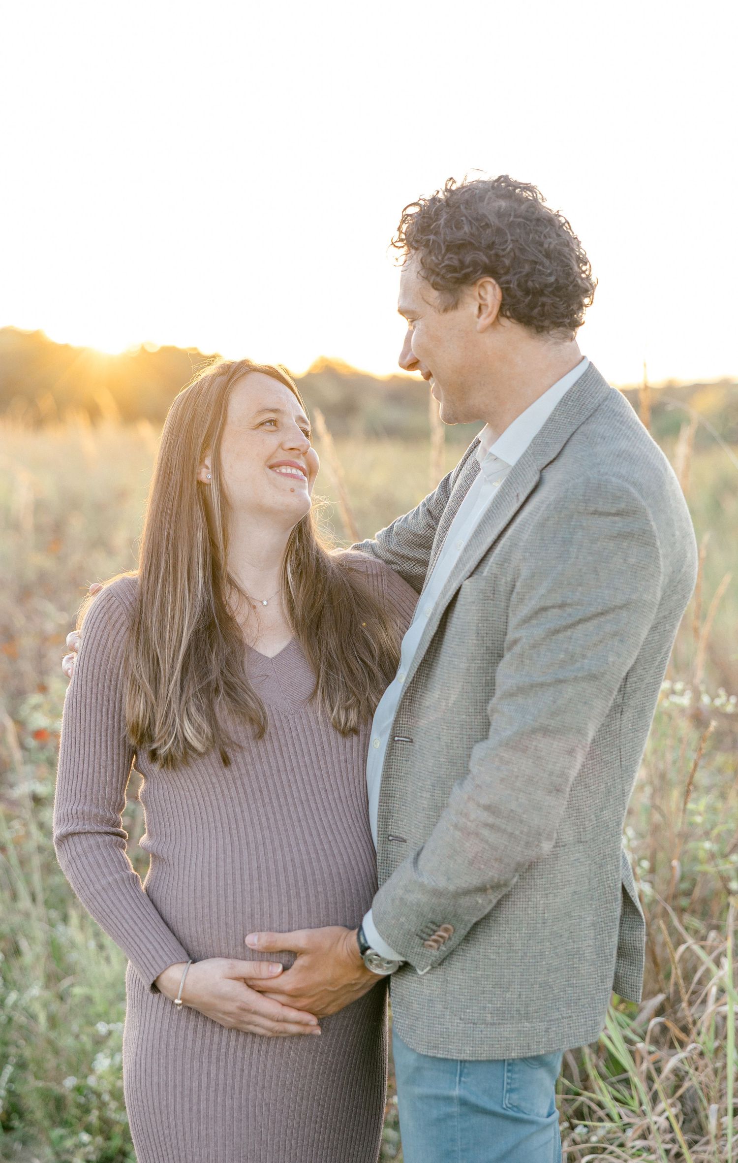 Couple smiling at each other in golden hour field, pregnant woman in mauve dress with partner in gray blazer.
