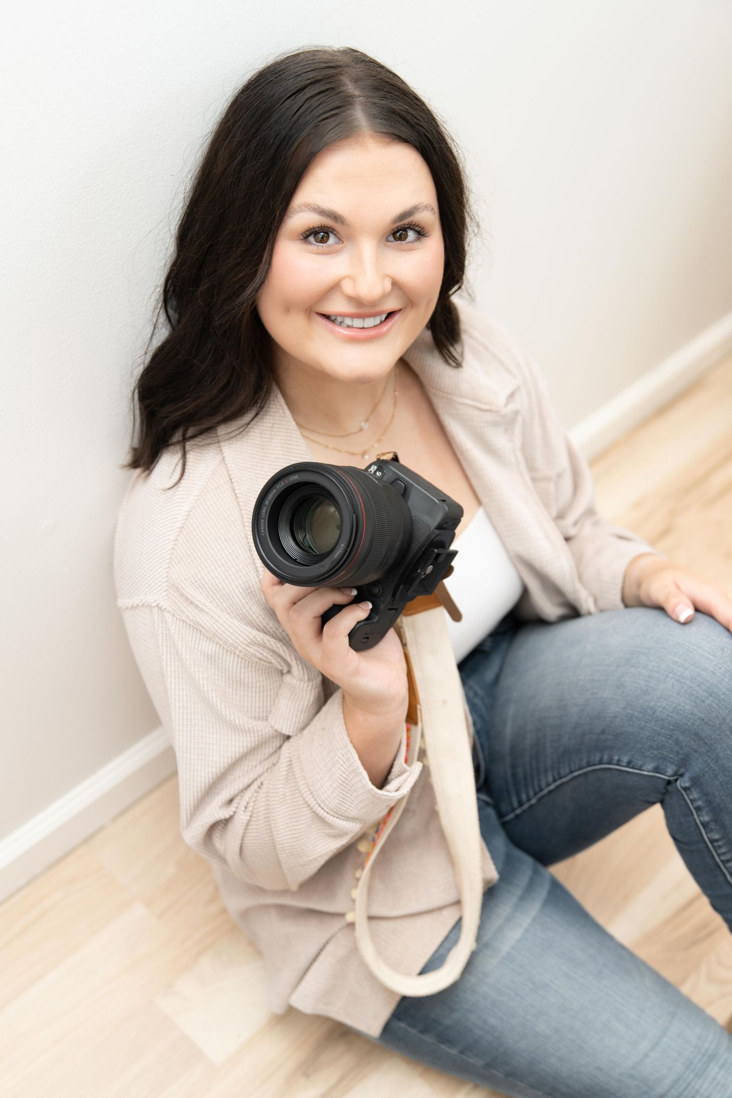 Smiling woman with dark hair sitting on floor holding a professional DSLR camera, wearing beige blazer and jeans.