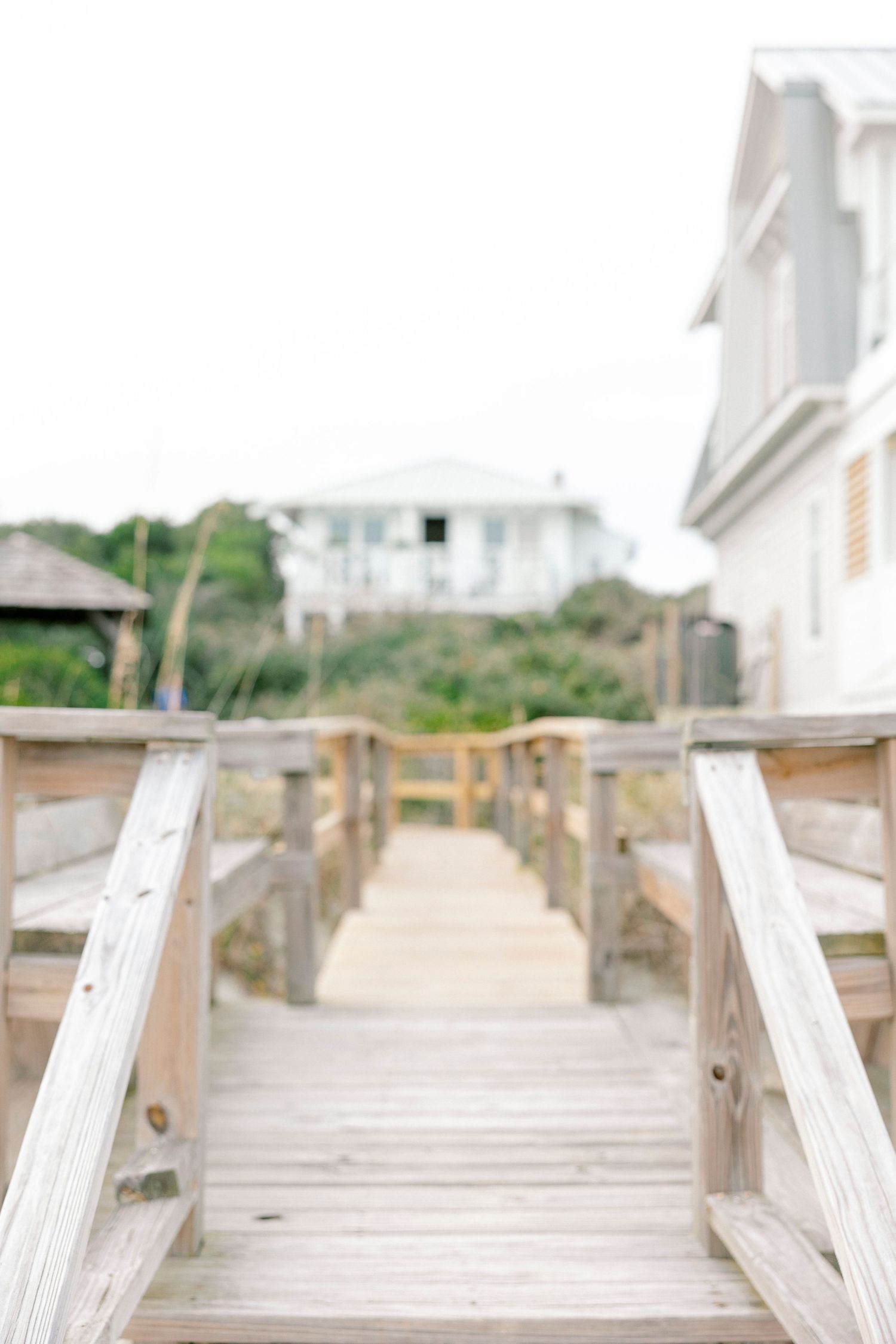 Wooden boardwalk leading to beach house with white railings and coastal scenery.