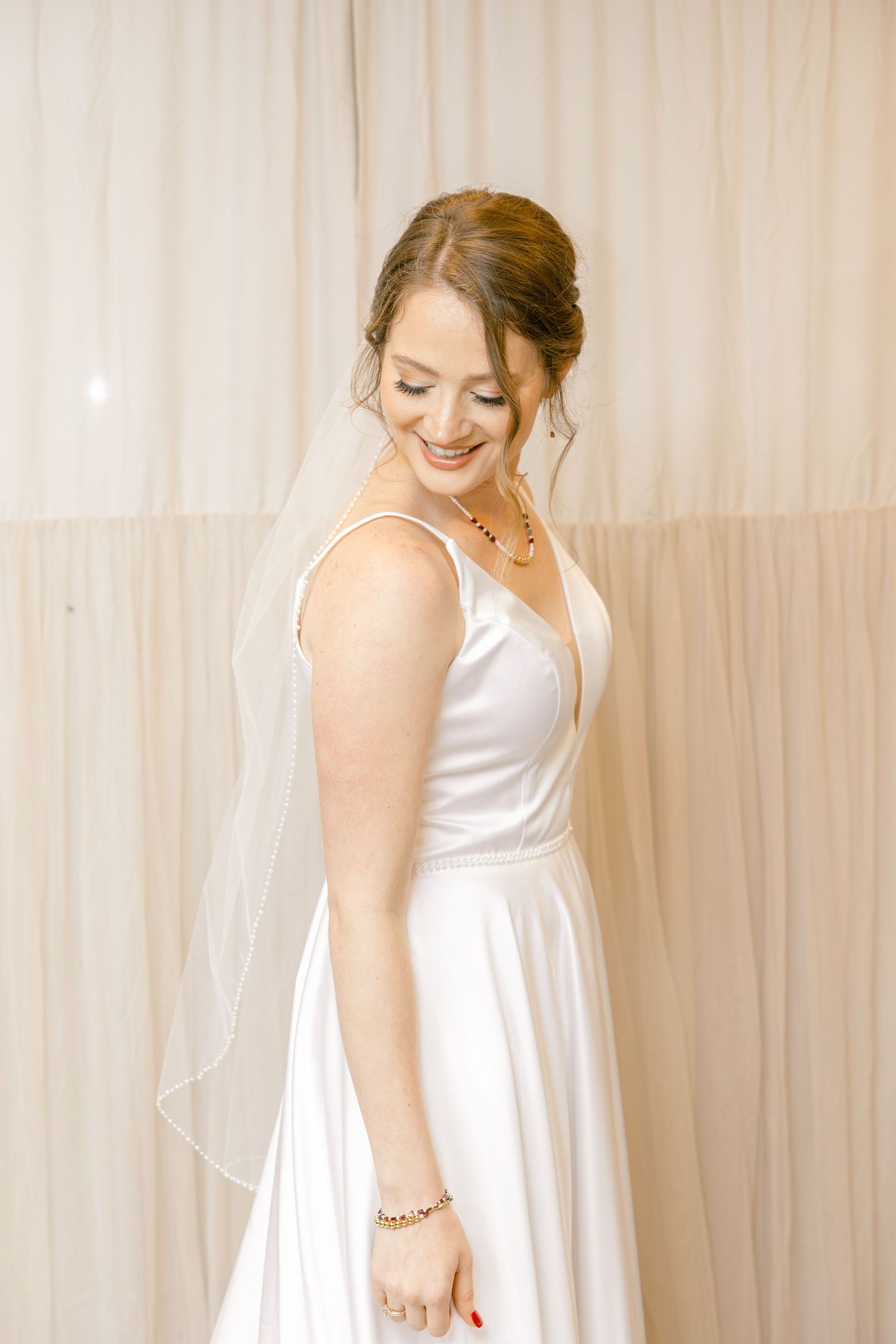 Smiling bride in white satin gown with veil and red necklace poses elegantly against soft draped backdrop.