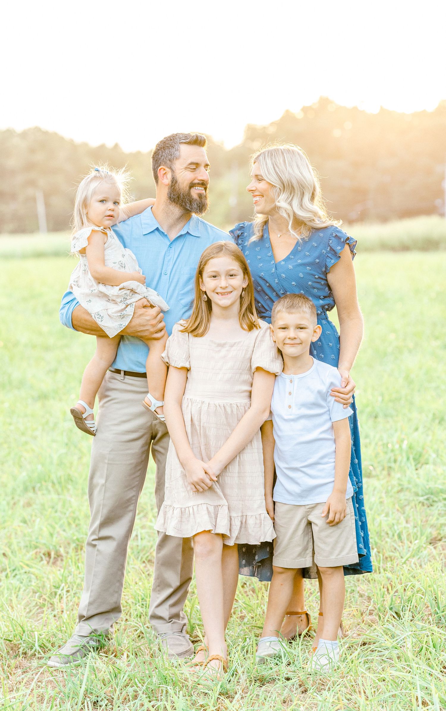 A happy family of five poses in a sunlit field, parents and three children smiling in coordinated outfits.