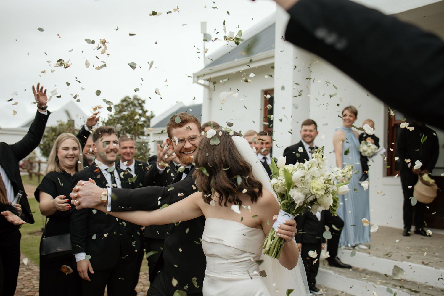 Wedding celebration with confetti being thrown outside a white building as newlyweds exit surrounded by joyful guests.