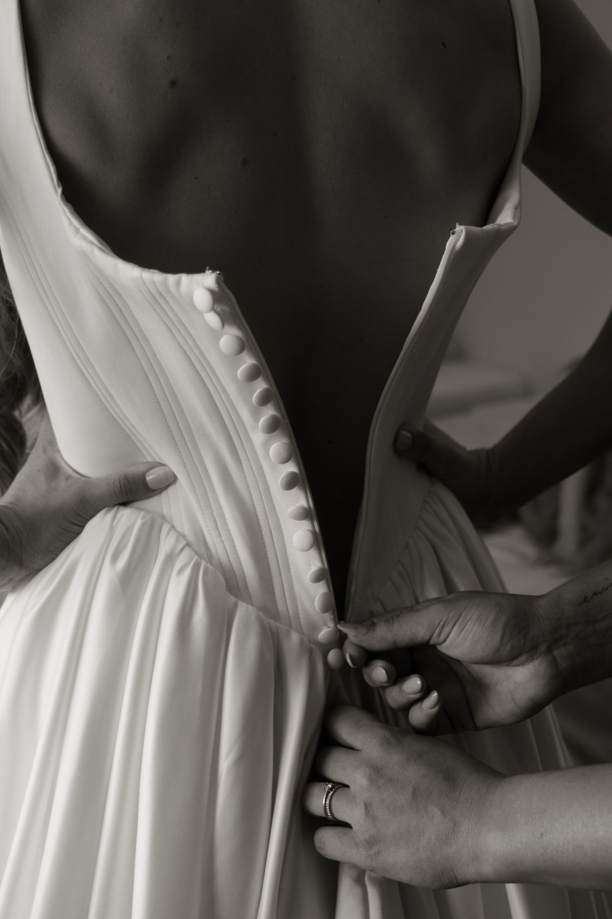Close-up black and white photo of hands fastening buttons on the back of a bridal gown.