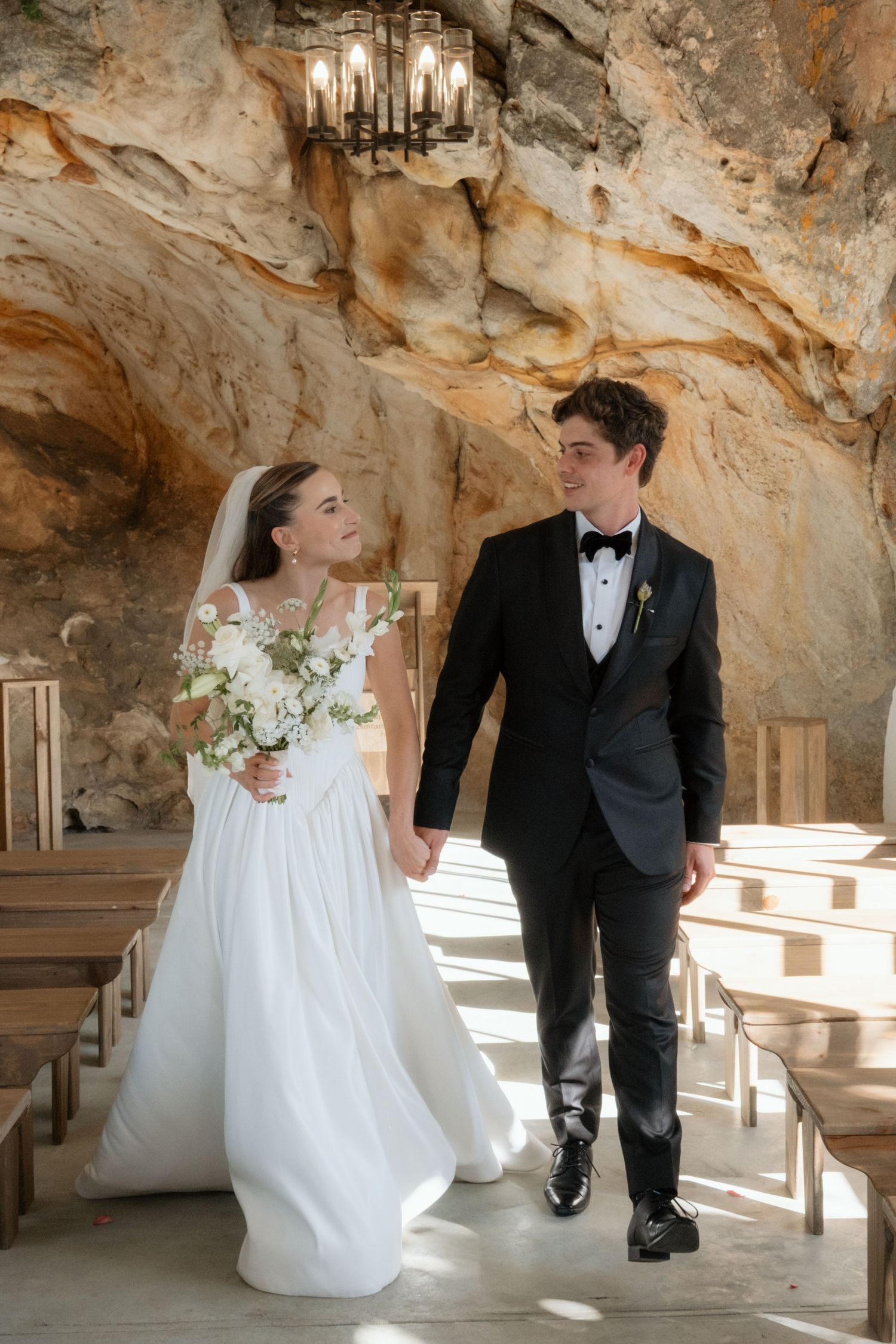 Bride and groom walking hand-in-hand through a stunning natural cave wedding chapel with wooden pews.