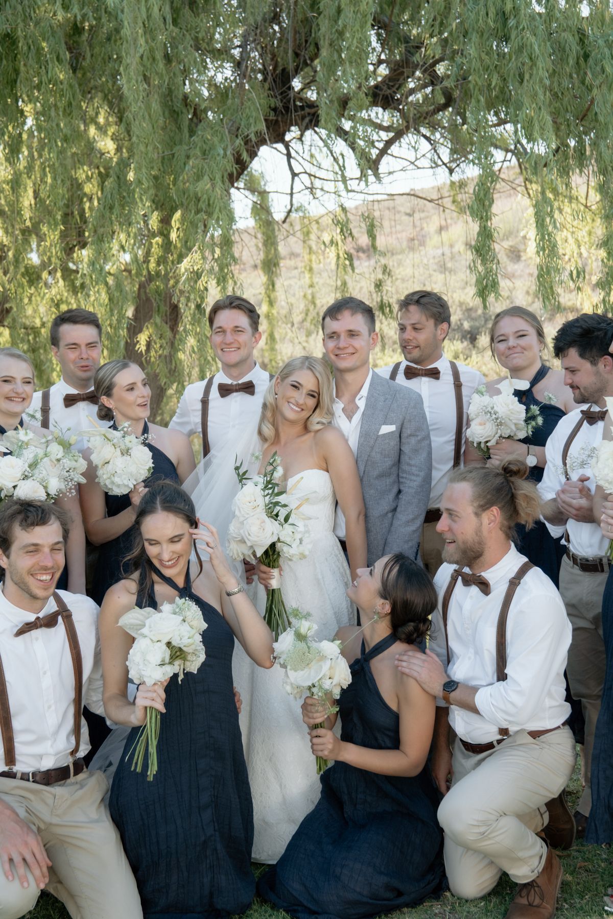Wedding party posing together under a willow tree, bridesmaids in navy dresses and groomsmen in suspenders.