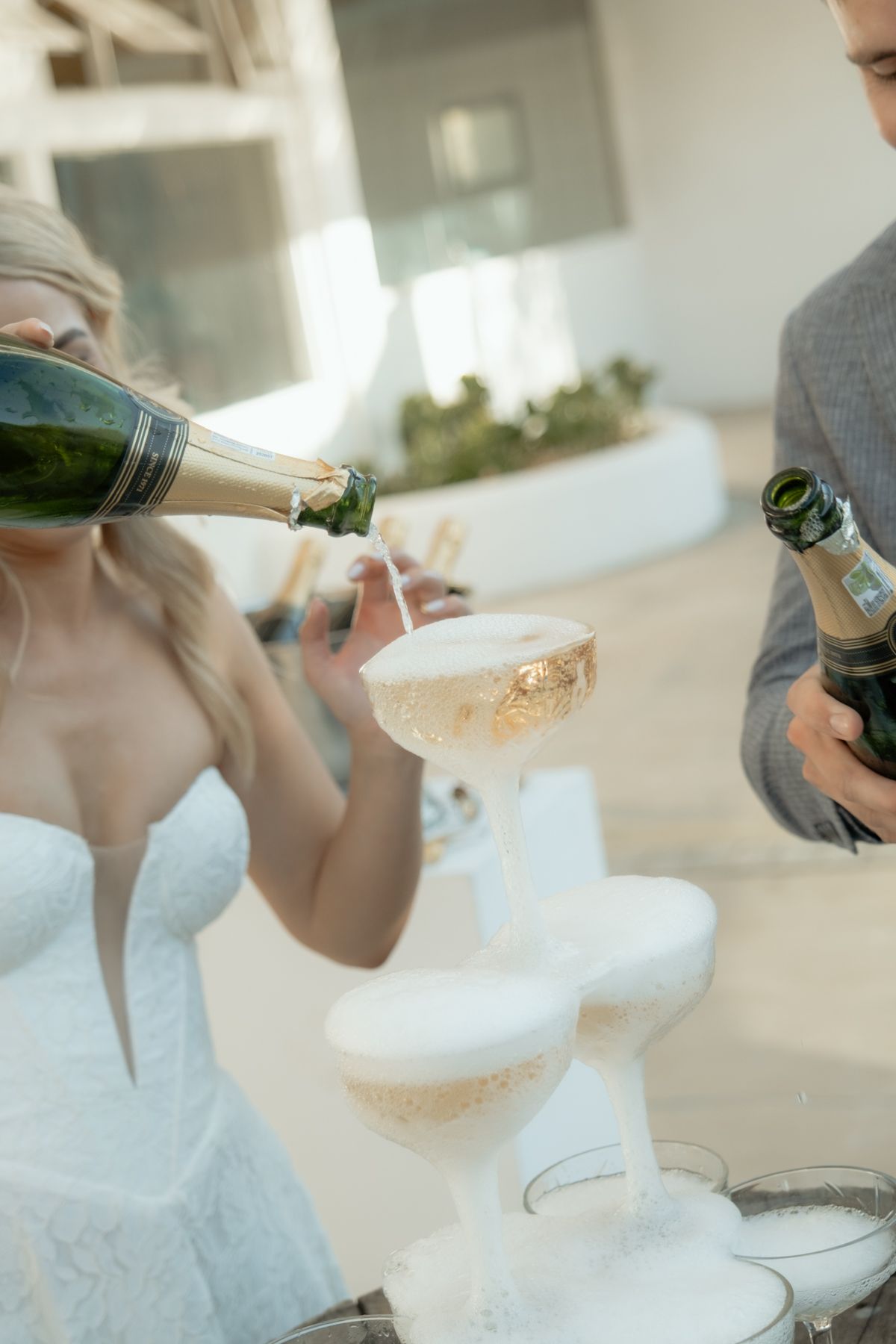 Bride pouring champagne into a tower of glasses at wedding reception while groom stands nearby.