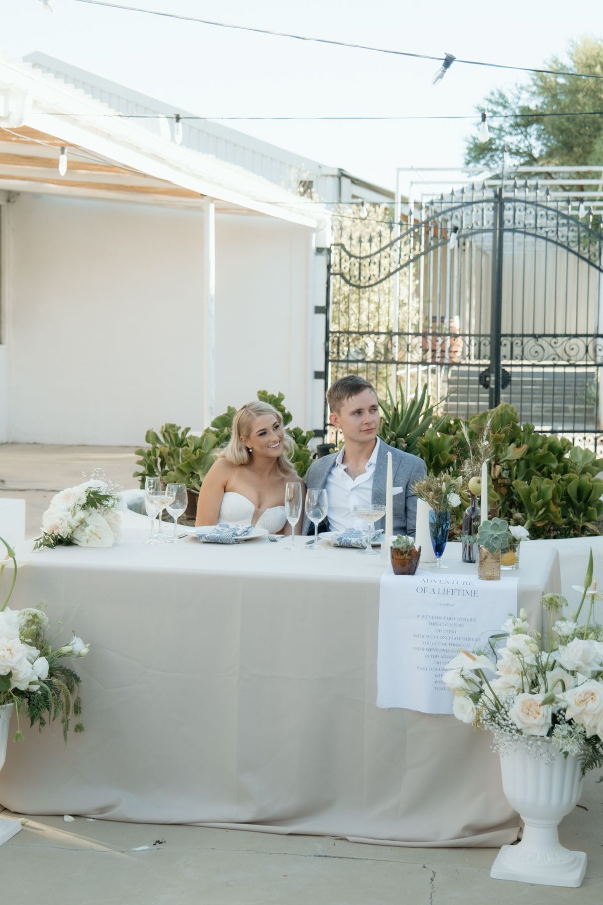 Bride and groom seated at a beautifully decorated sweetheart table with white floral arrangements at their outdoor reception.