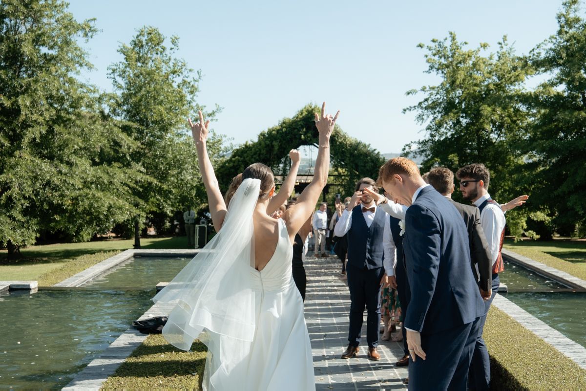 Joyful bride raises her hands in celebration while walking with groom and wedding party beside a garden fountain.