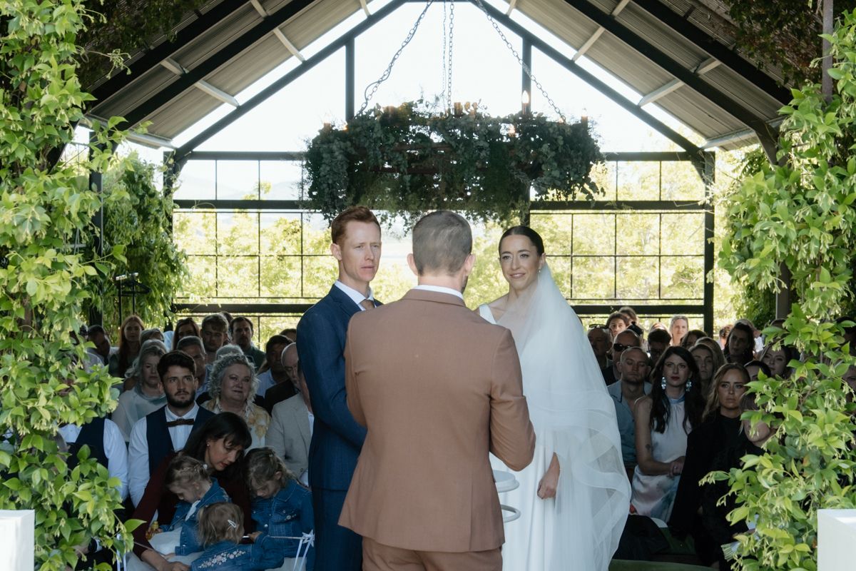 Bride and groom exchanging vows in a glass chapel adorned with lush greenery and guests seated.
