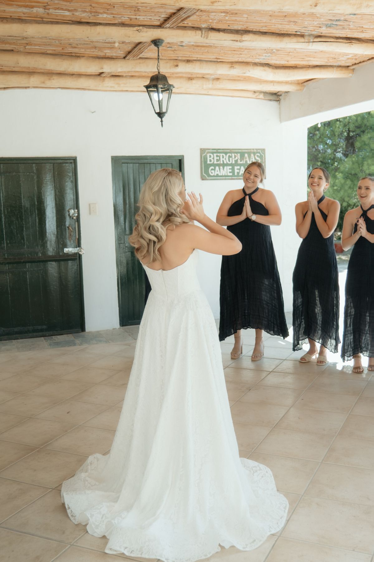Bride in a strapless white lace gown reveals herself to bridesmaids dressed in black at a wedding.