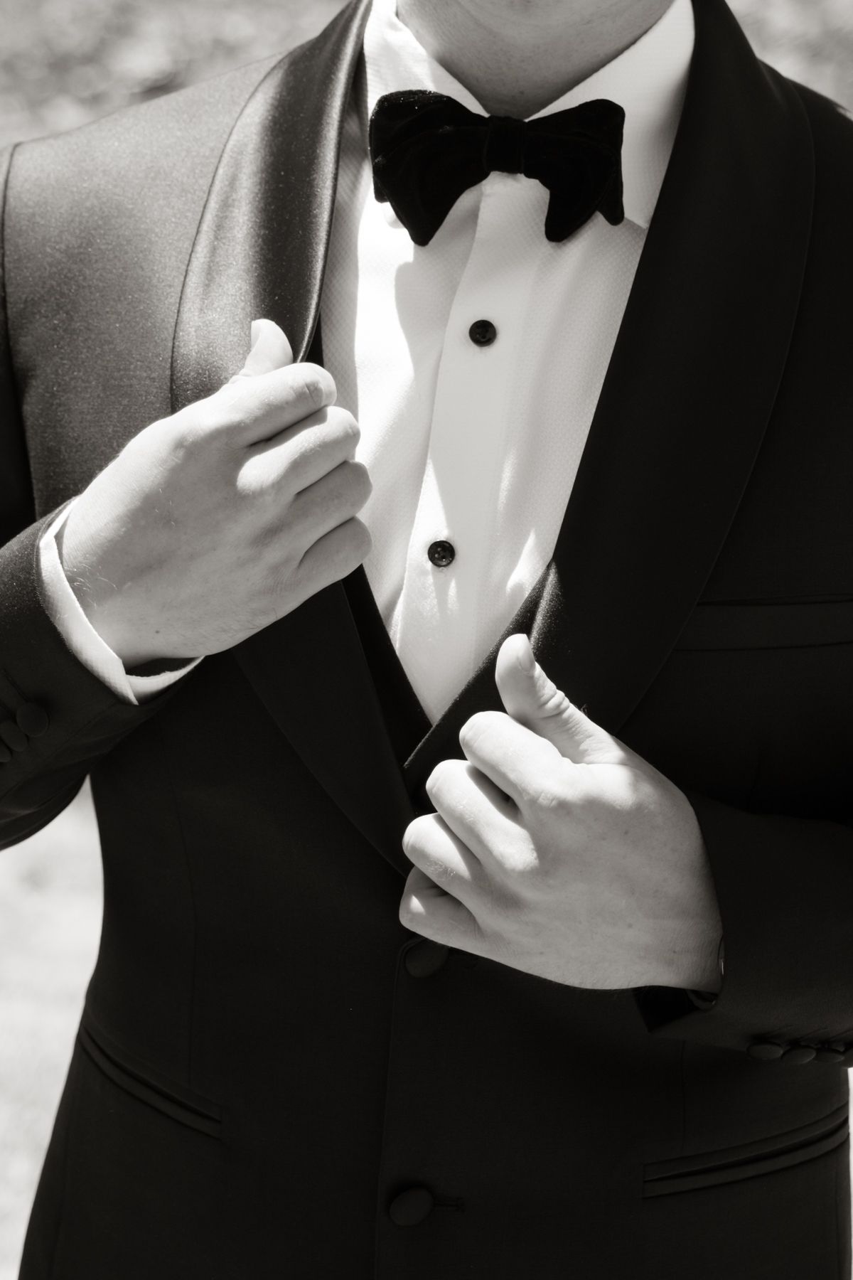 Black and white close-up of groom adjusting elegant black tuxedo with velvet bow tie.
