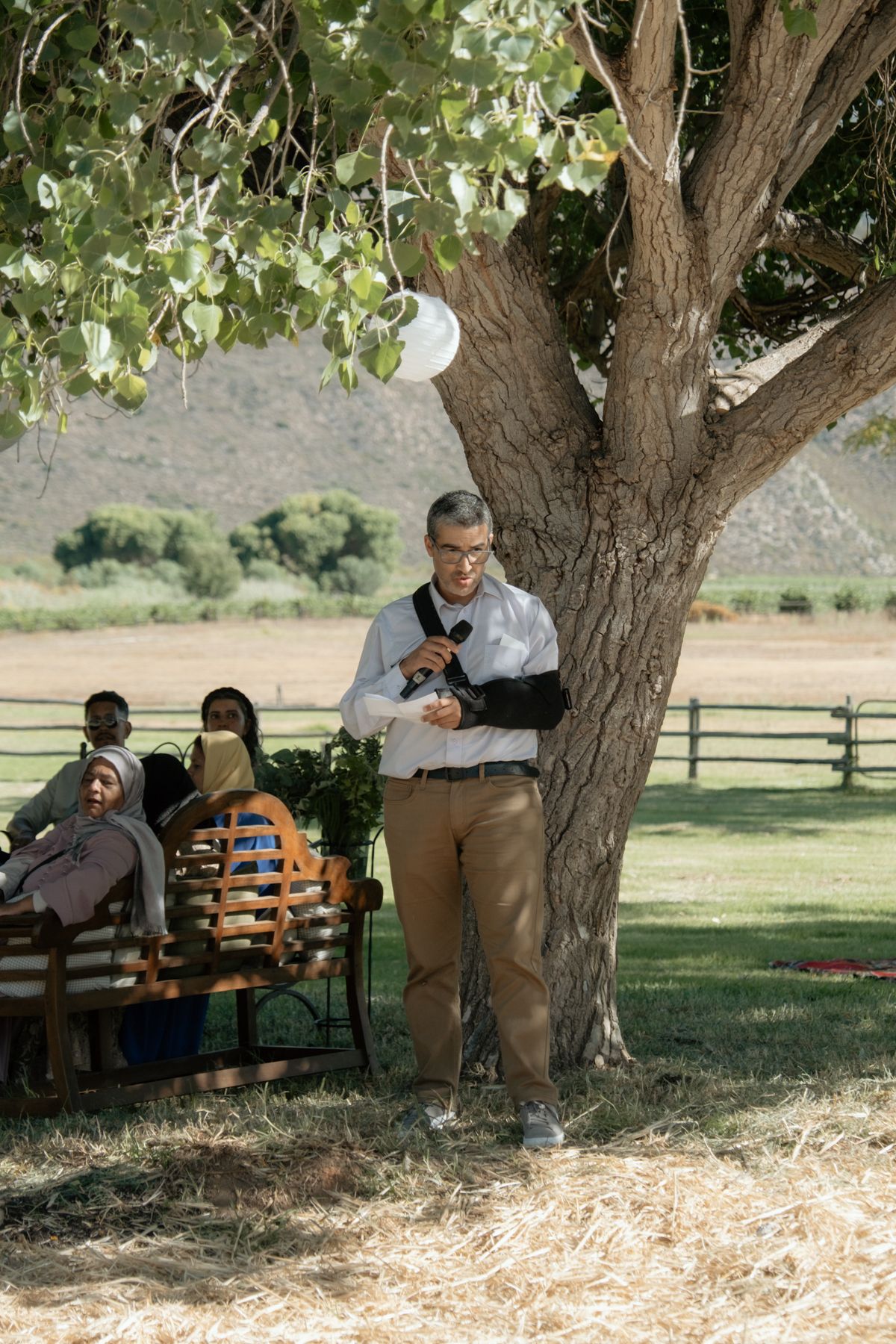 Wedding officiant standing under a large tree reading from notes at an outdoor farm wedding ceremony.