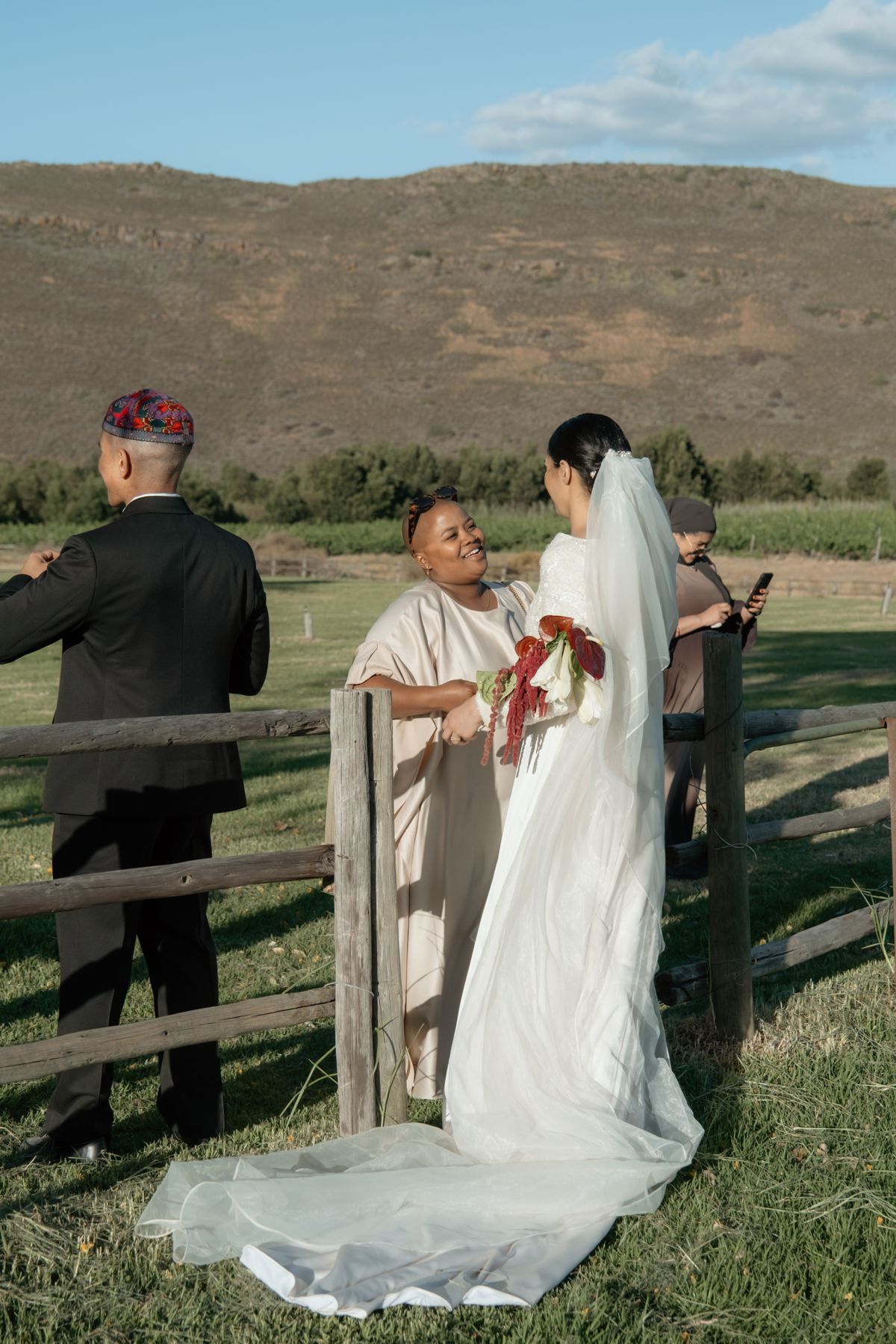 Bride in white gown with long veil chatting with guest by rustic wooden fence at farm wedding.