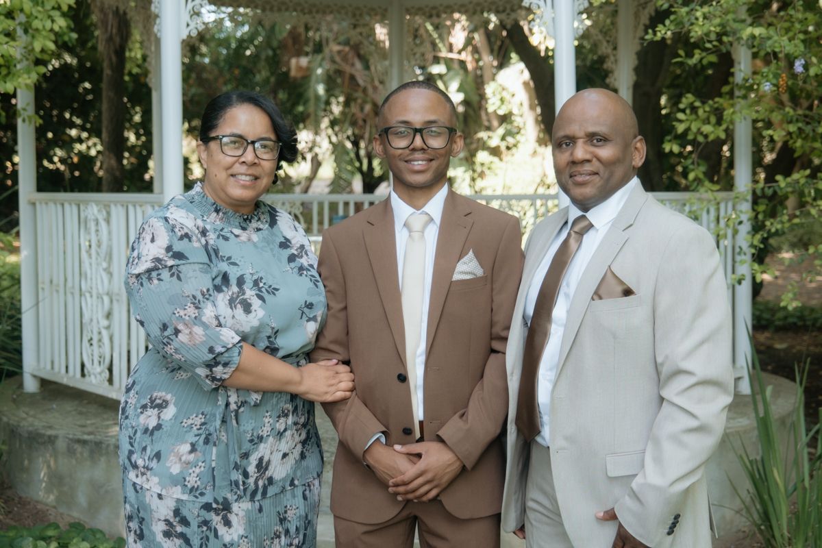 Smiling groom in brown suit stands between proud parents near white garden gazebo outdoors.