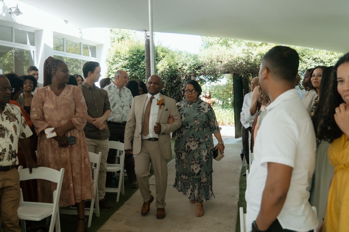 Guests gather under a white tent for an outdoor wedding ceremony, with the wedding party walking down the aisle.