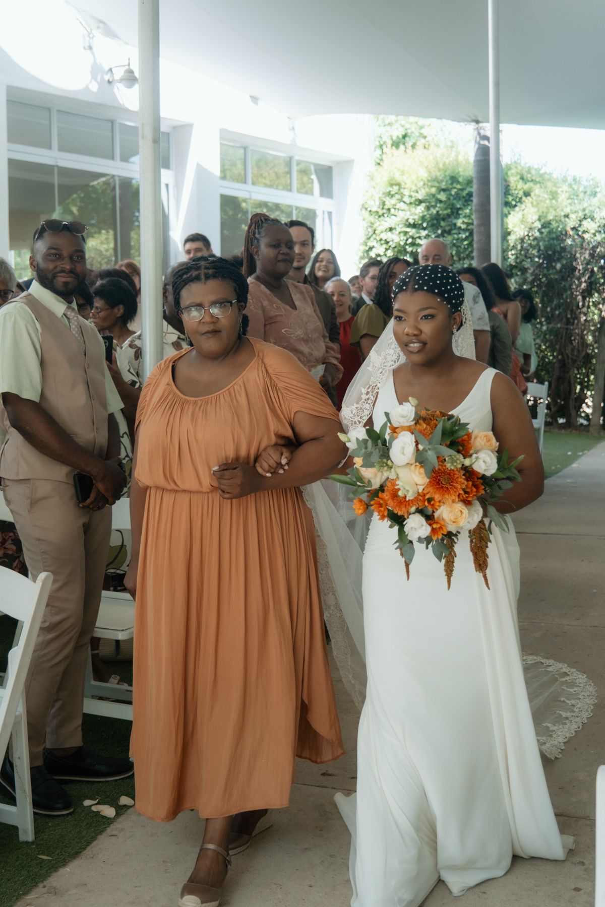 Bride in white gown holding orange and white bouquet walks down aisle with woman in orange dress.
