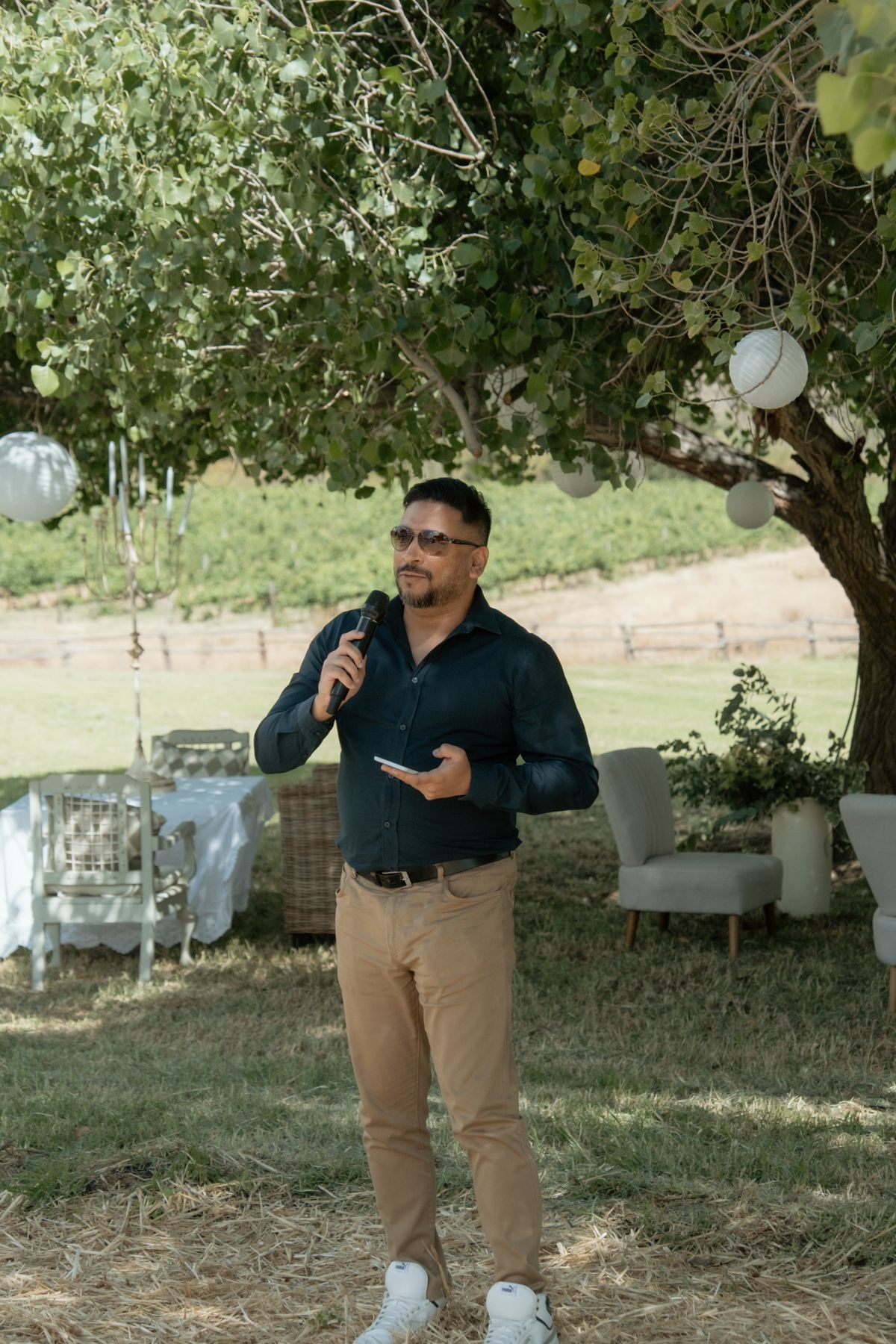 Man in dark shirt speaks into microphone at outdoor wedding reception under large tree.