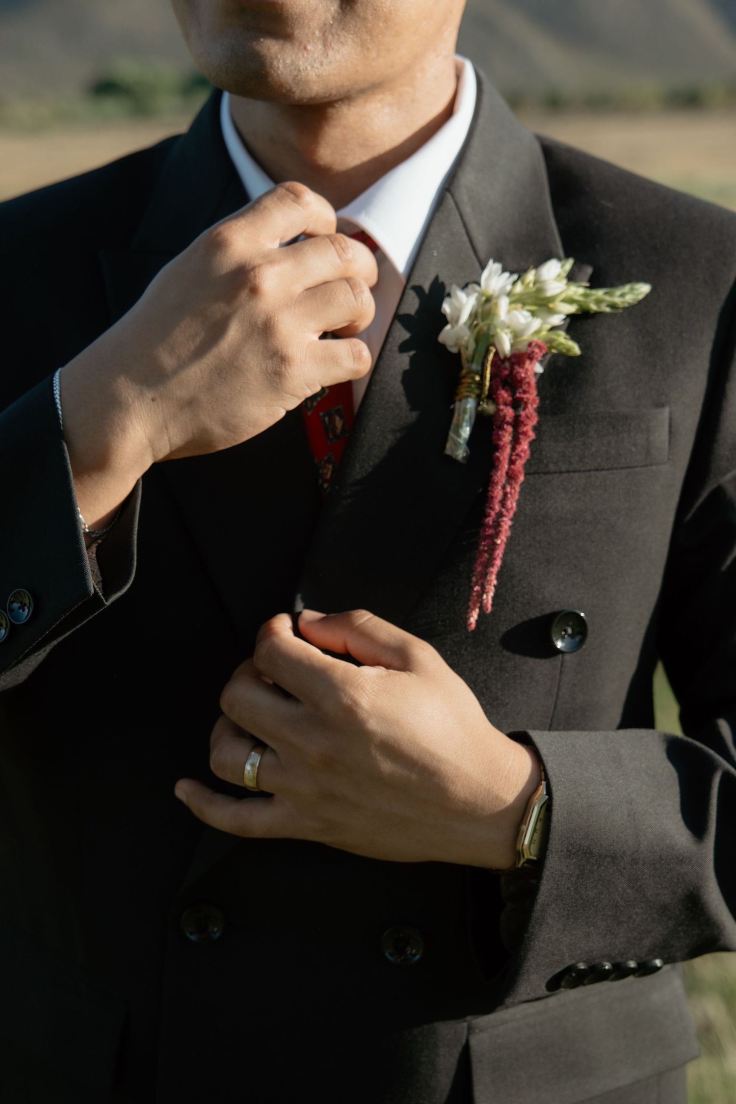 Groom in black suit adjusts boutonniere with white and red flowers at outdoor wedding.