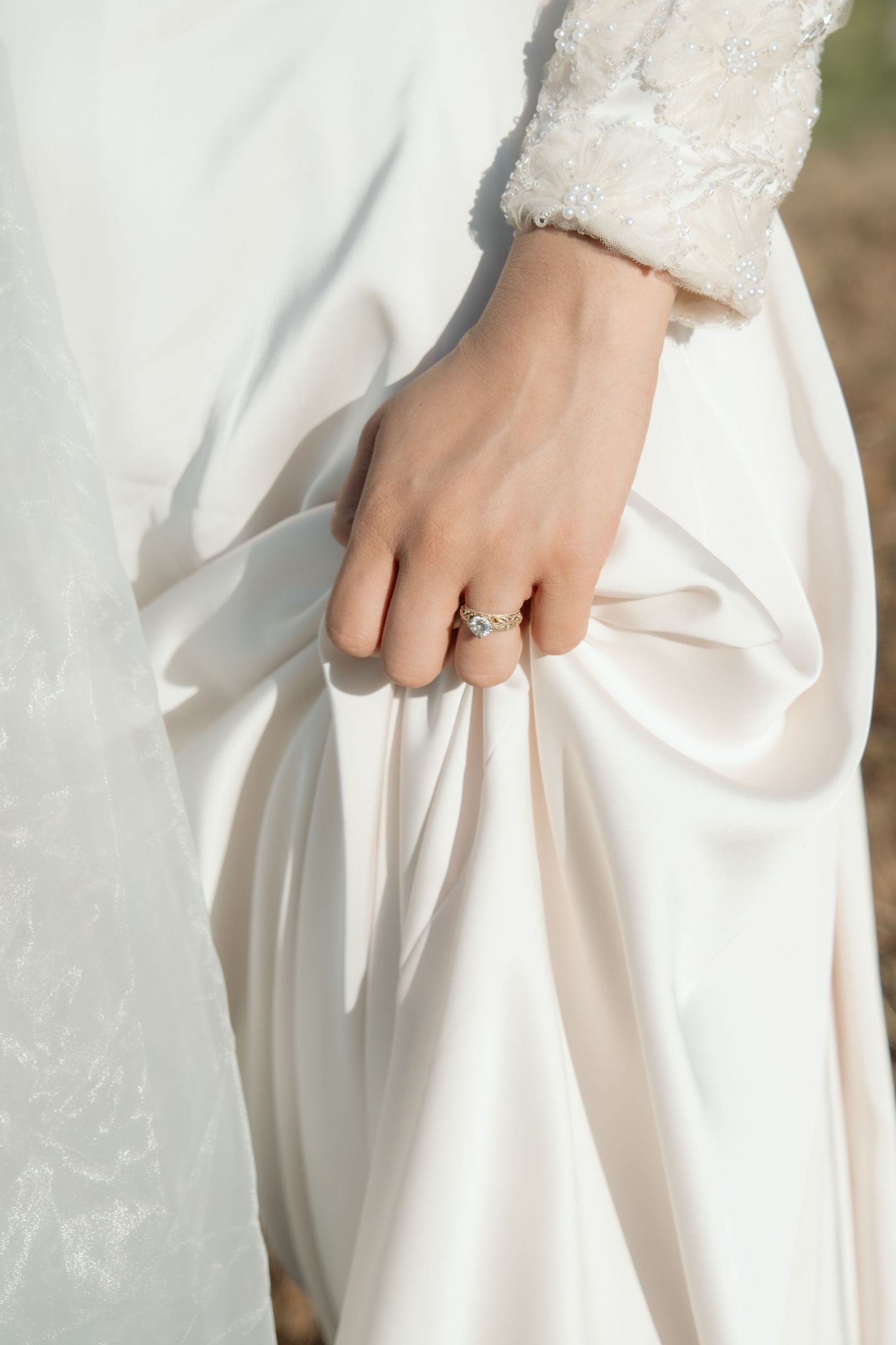 Close-up of a bride's hand holding white satin wedding dress, wearing a diamond engagement ring.
