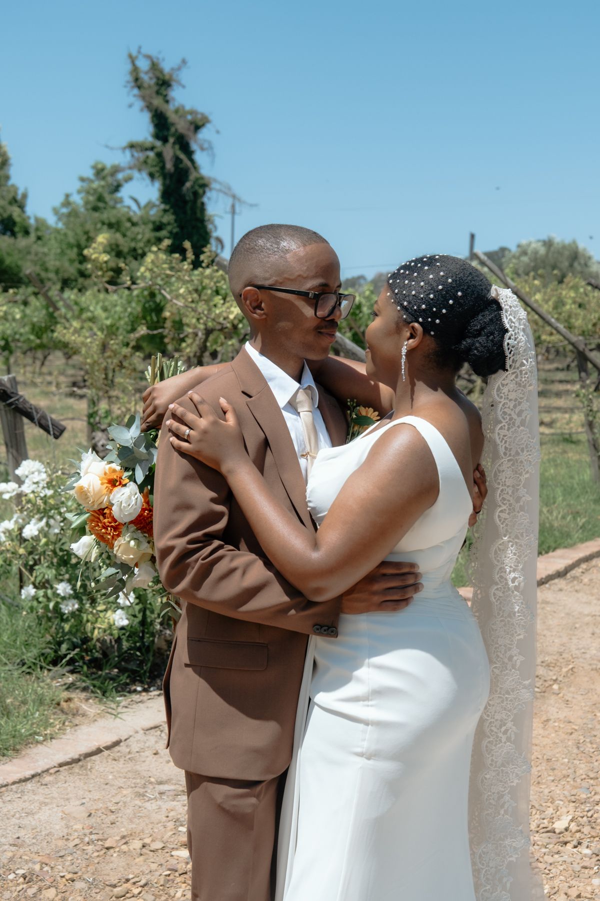 A Black couple shares a romantic moment in a vineyard, the groom in a brown suit and bride in a white gown with veil.