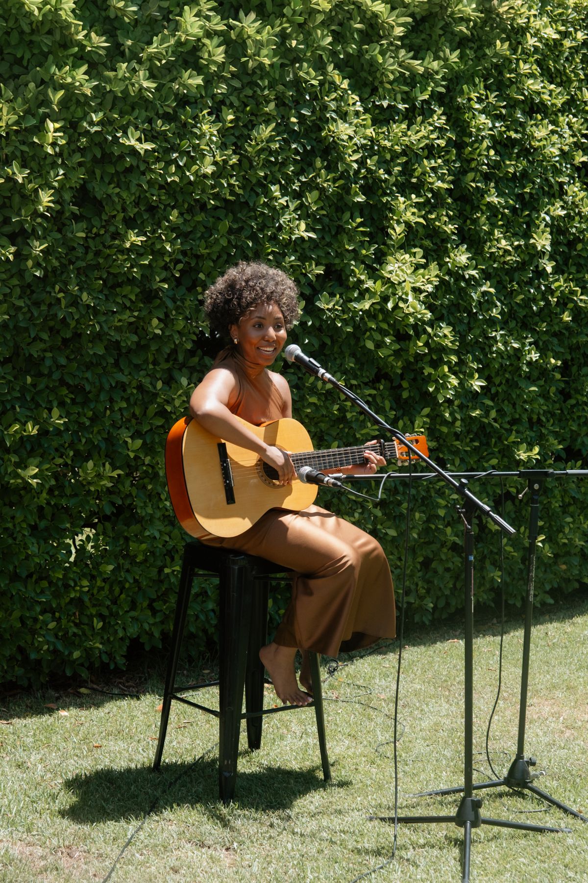 Woman playing acoustic guitar and singing into a microphone outdoors, seated on a stool against a green hedge backdrop.