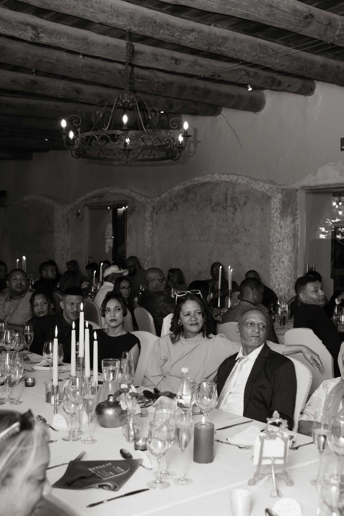 Wedding guests seated at candlelit reception tables in rustic venue with wooden beam ceiling.