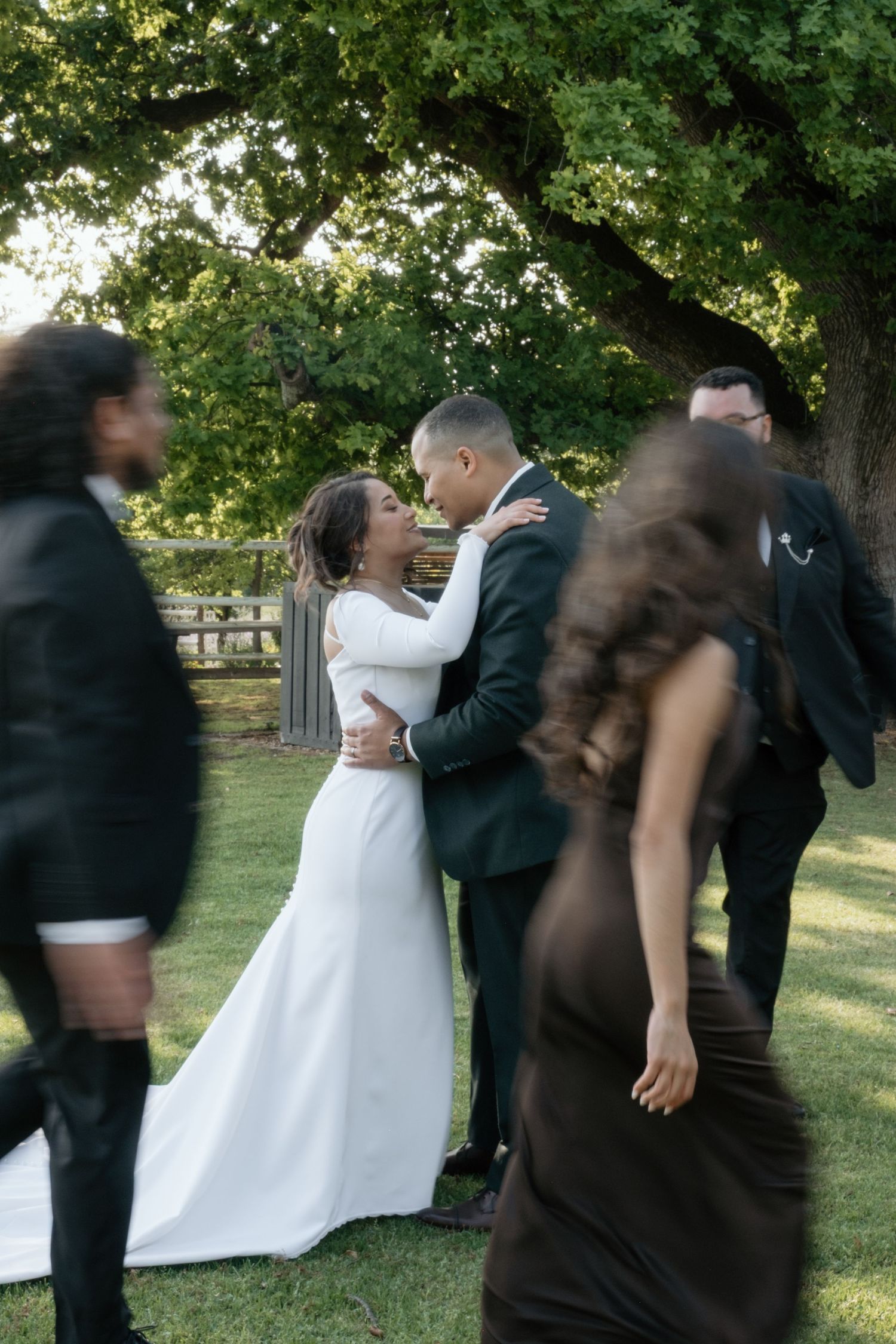 Bride and groom share a romantic moment outdoors under a large tree while wedding guests move around them.