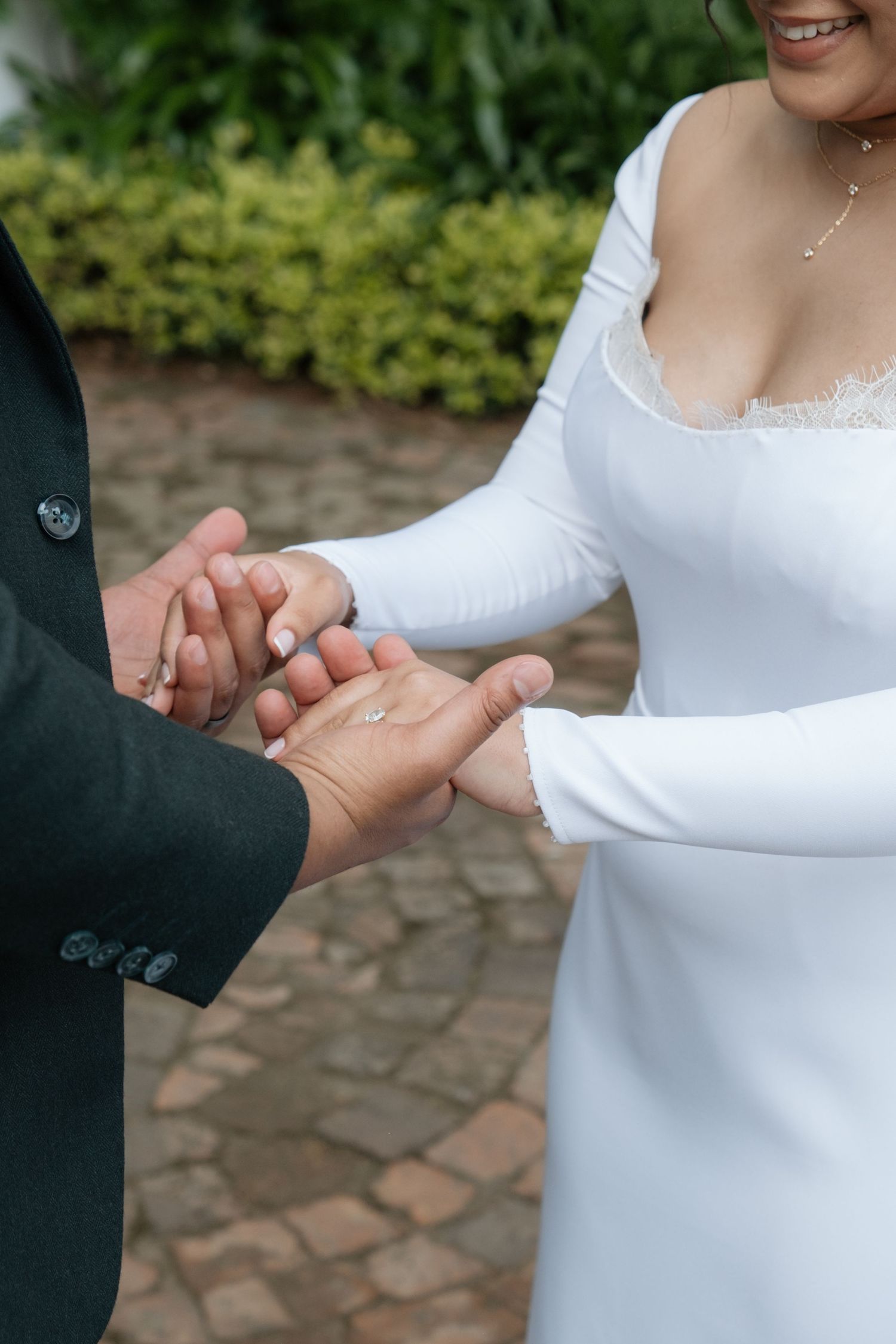 Bride and groom exchange rings outdoors, with bride in elegant long-sleeve white wedding gown on cobblestone path.