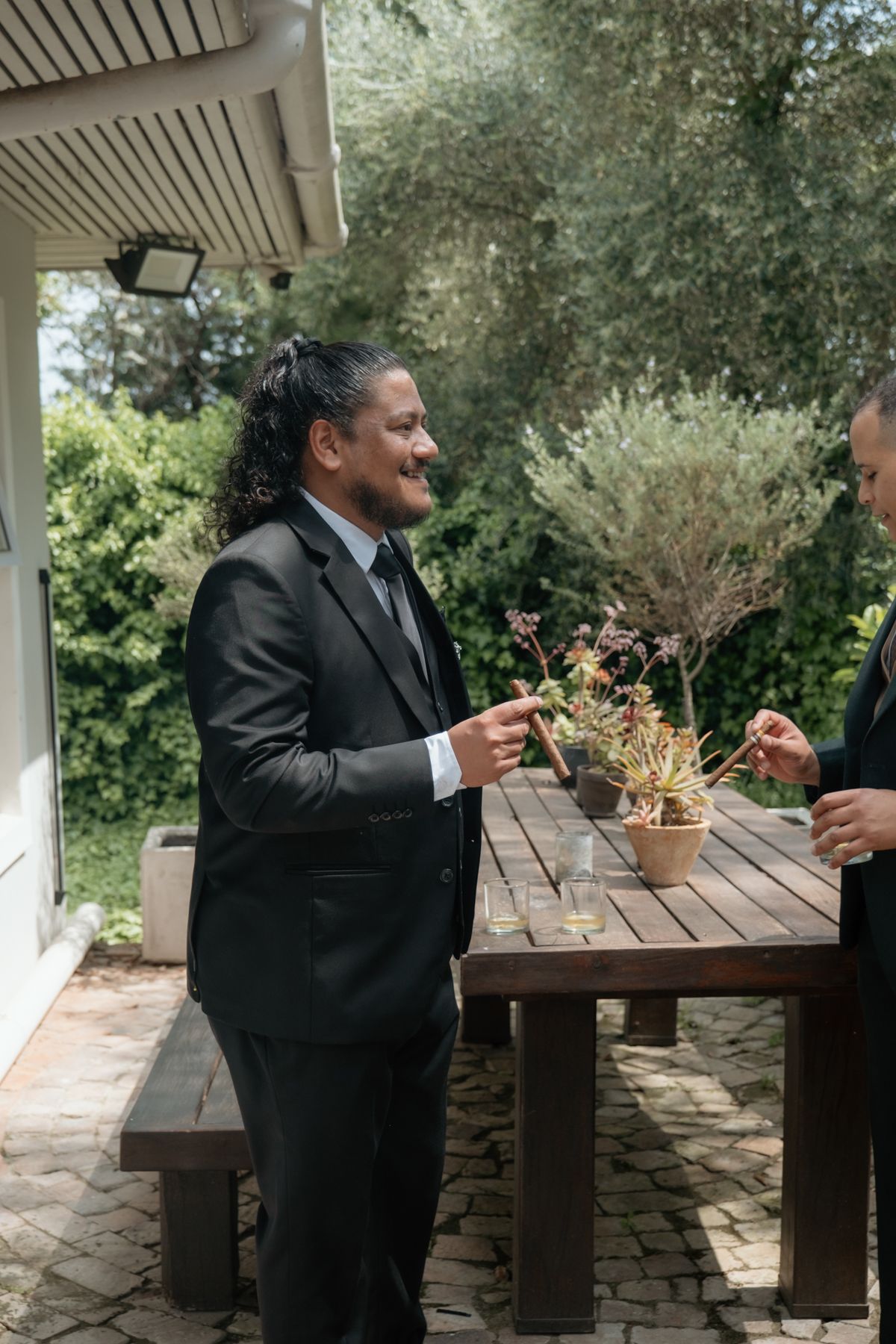 Smiling man with long hair in black suit laughing at outdoor wedding reception patio table.