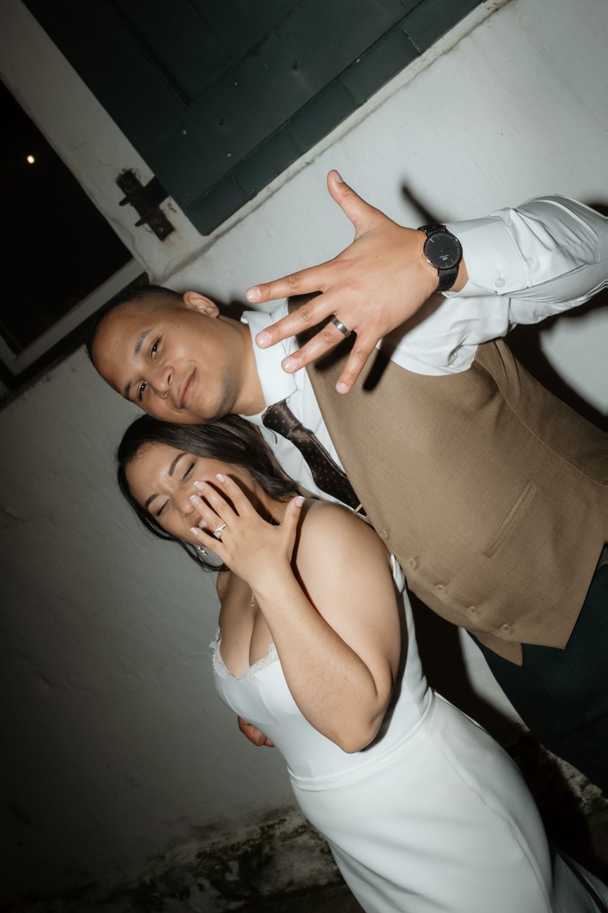 Couple posing playfully showing off wedding rings, groom in vest and bride in white strapless gown.