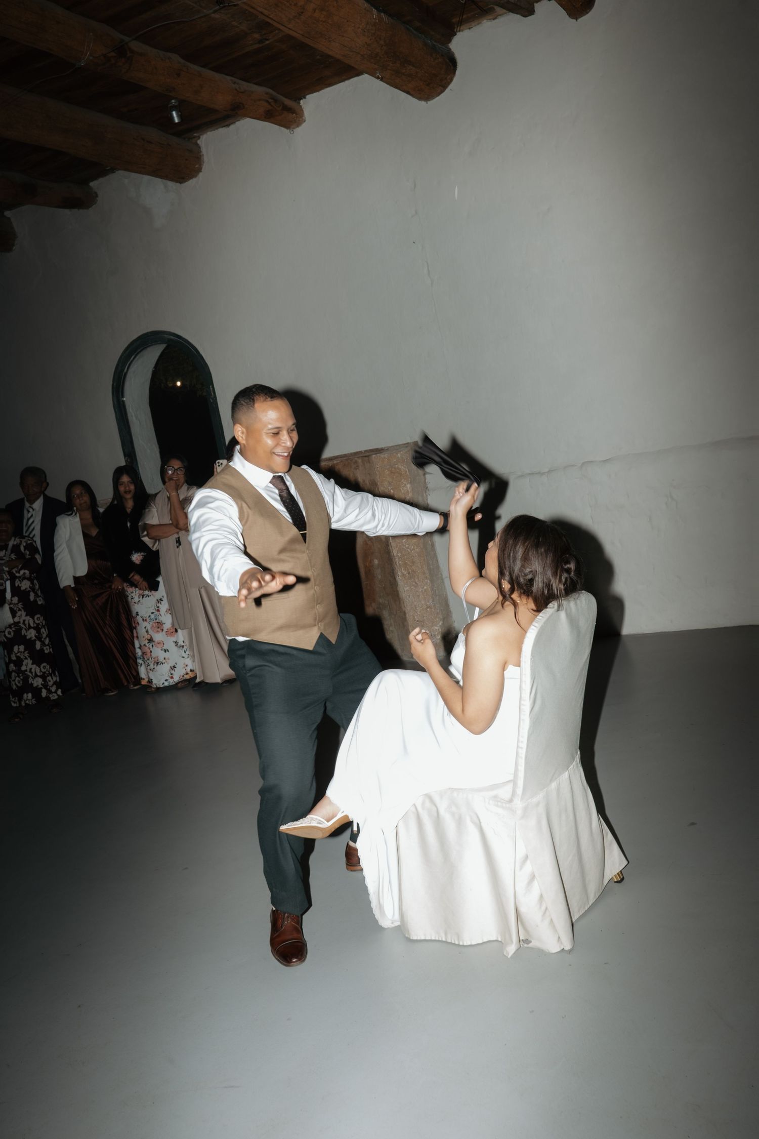 Groom dances playfully with seated bride during fun wedding reception shoe game in whitewashed rustic venue.