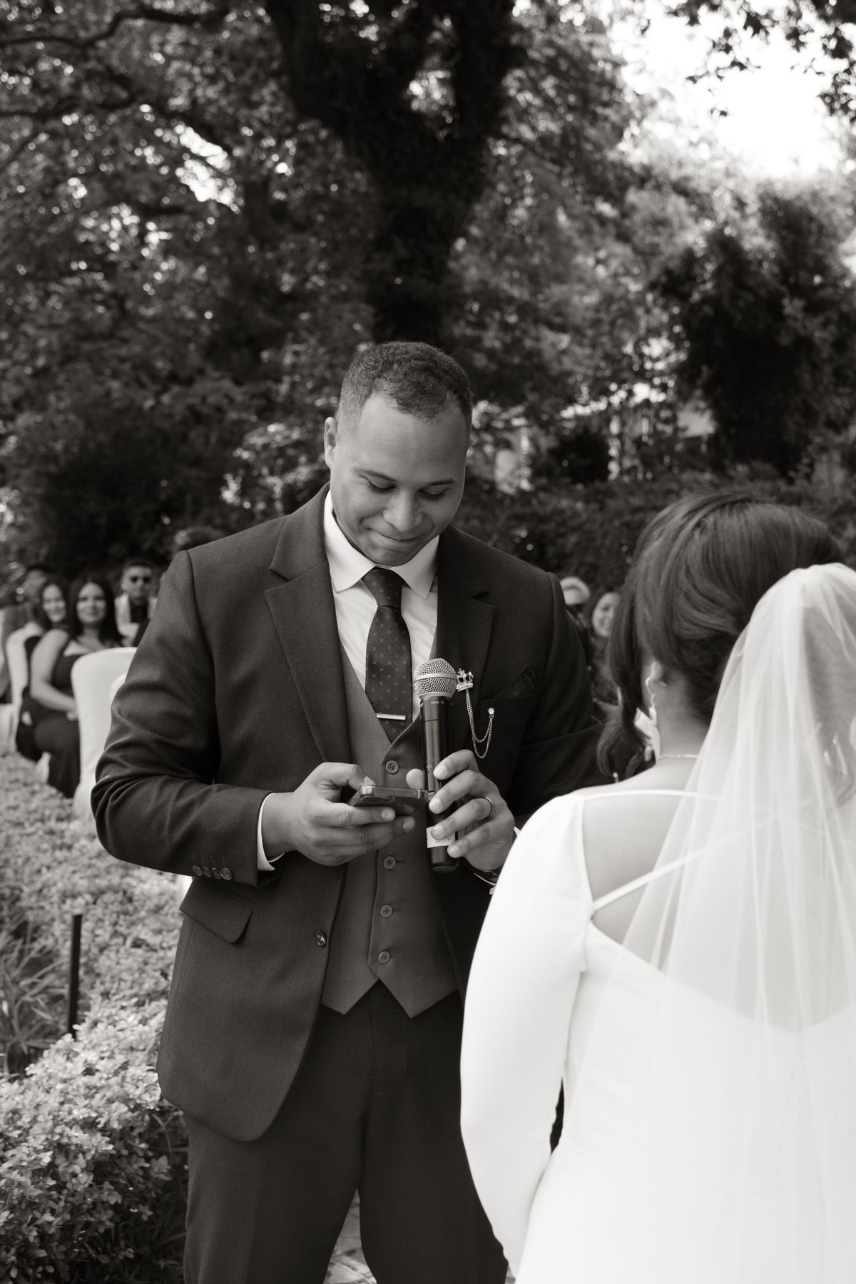 Groom reading vows from phone during outdoor wedding ceremony while bride in veil stands opposite in black and white photo.