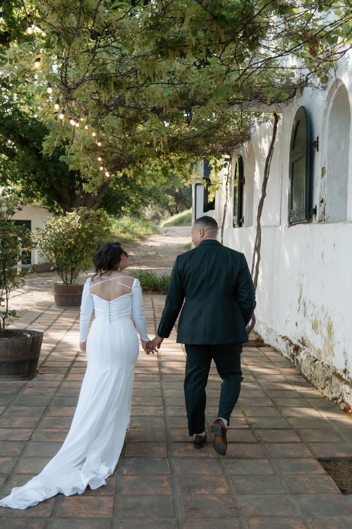 A bride and groom walk hand in hand along a white-walled garden path at their venue.