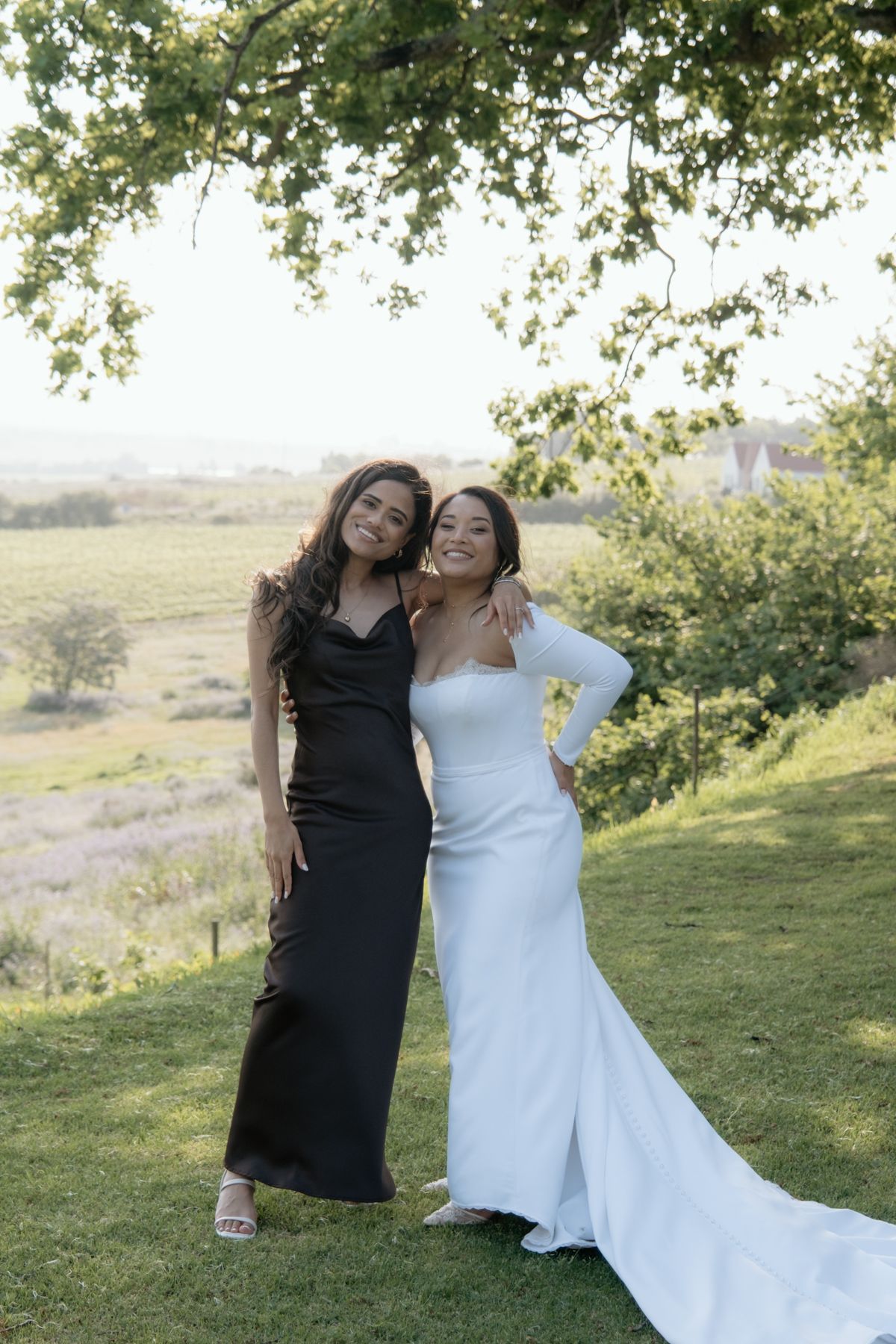 Bride in white gown poses with guest in black dress under tree at outdoor vineyard wedding.