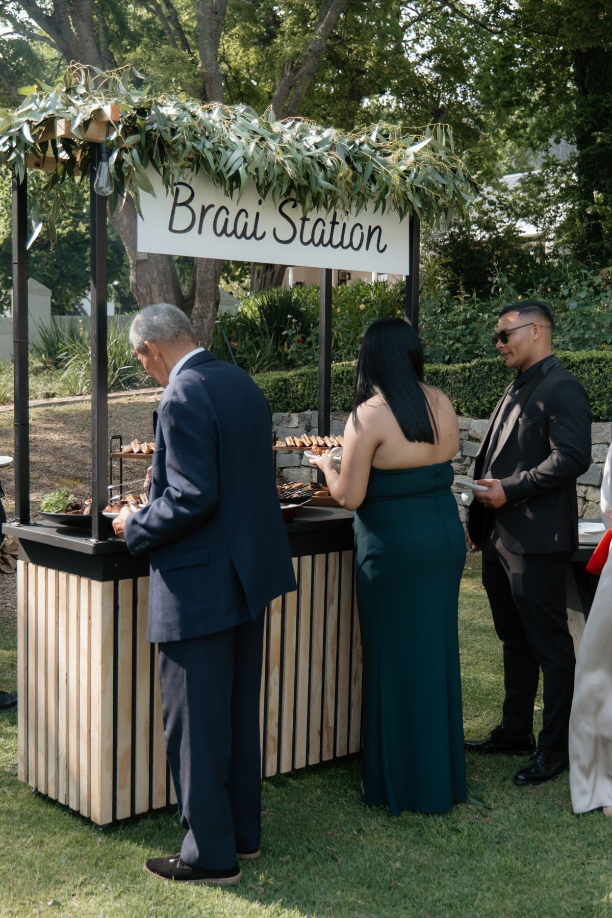 Guests serving themselves at an elegant outdoor Braai Station decorated with greenery at a wedding reception.
