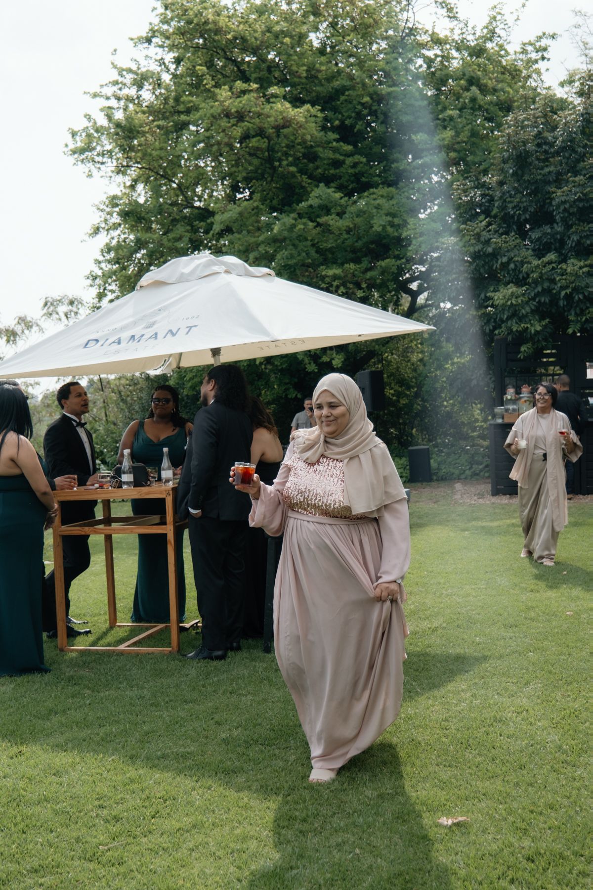 A woman in a beige hijab and long dress holds a drink at an outdoor wedding reception under a white umbrella.