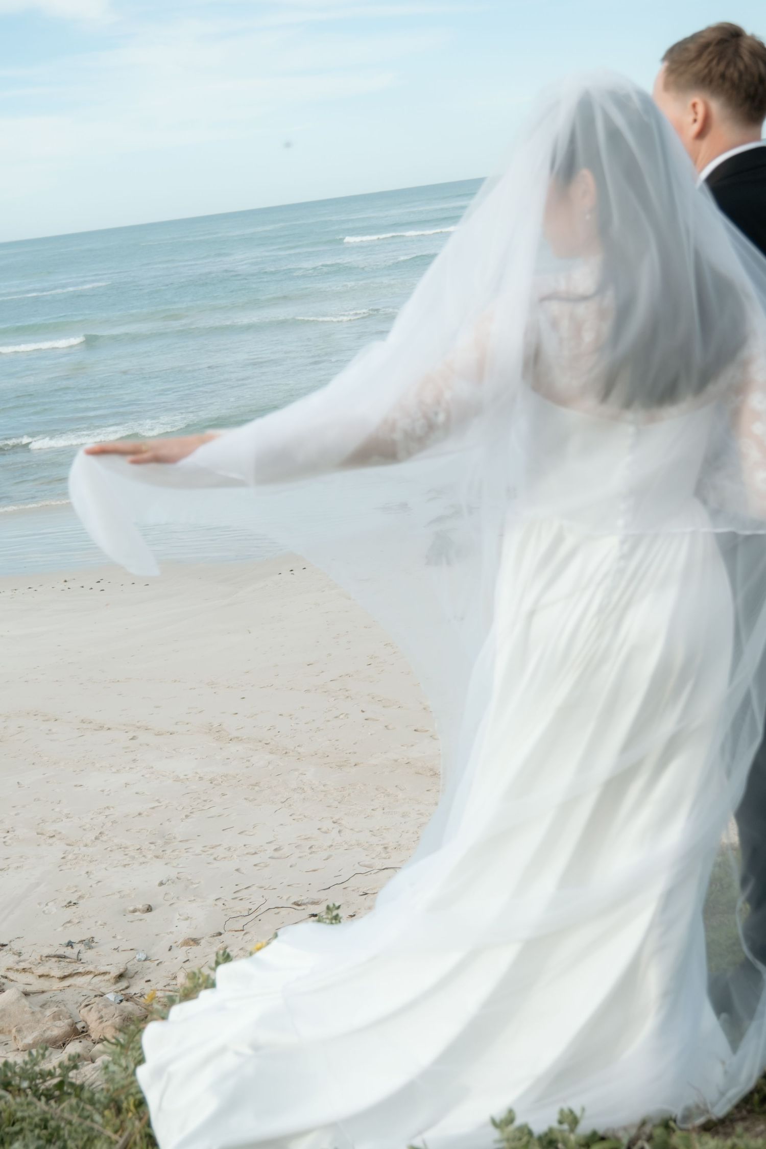 Bride in flowing white gown and veil stands with groom overlooking scenic ocean beach.