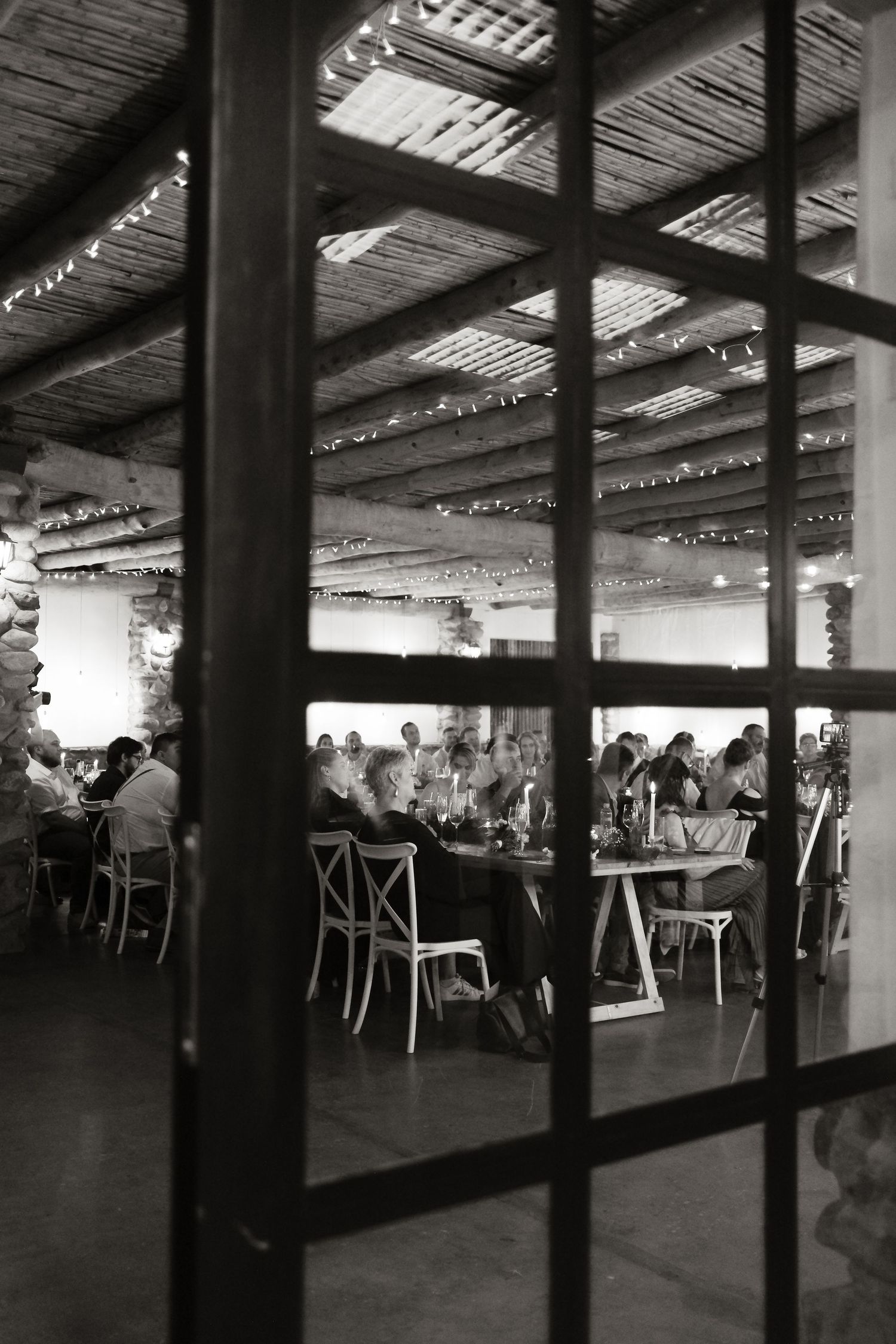 Black and white photo of diners gathered at tables inside a rustic restaurant viewed through glass doors.