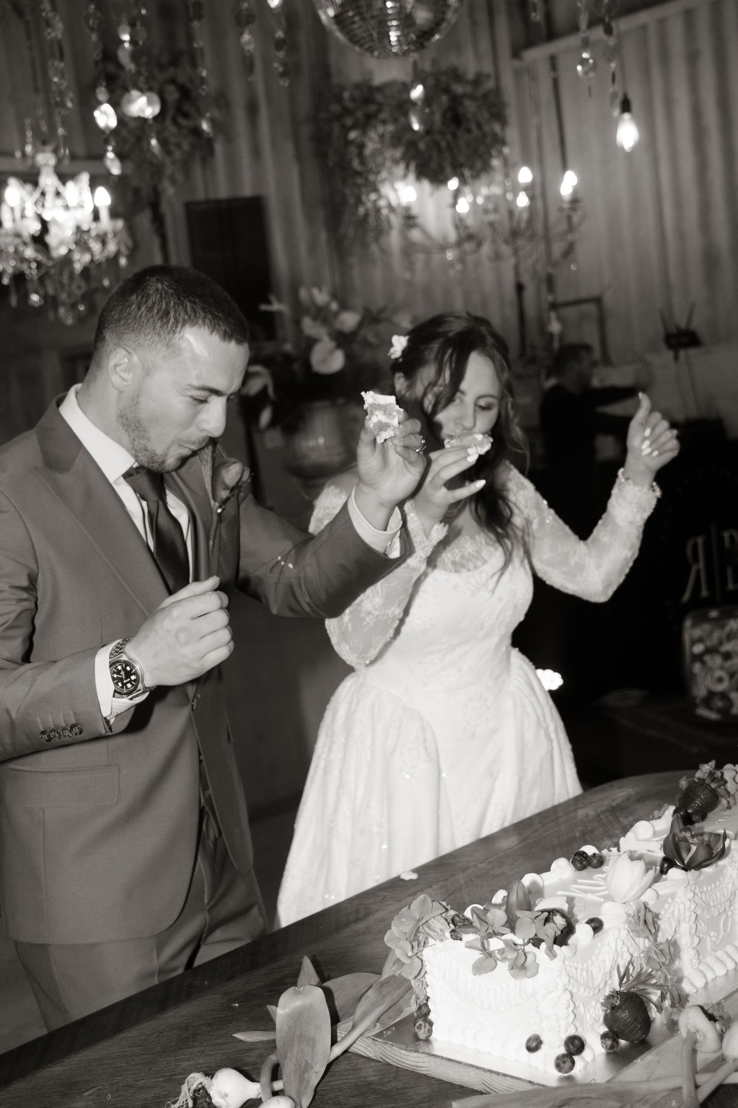 Bride and groom playfully feed each other wedding cake at elegant chandelier-lit reception in black and white.