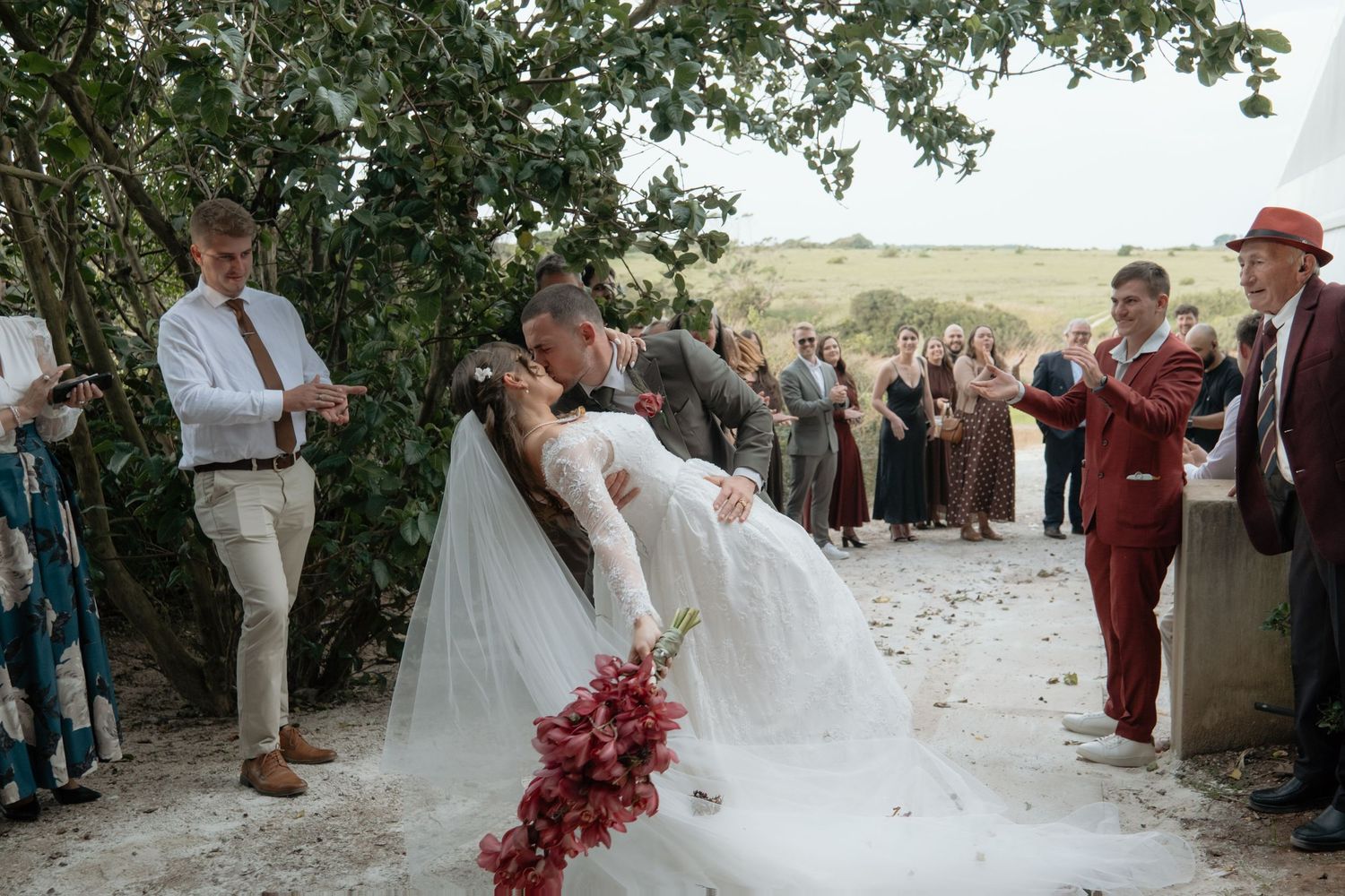 Bride and groom share a romantic dip kiss at outdoor wedding ceremony while guests clap and celebrate around them.