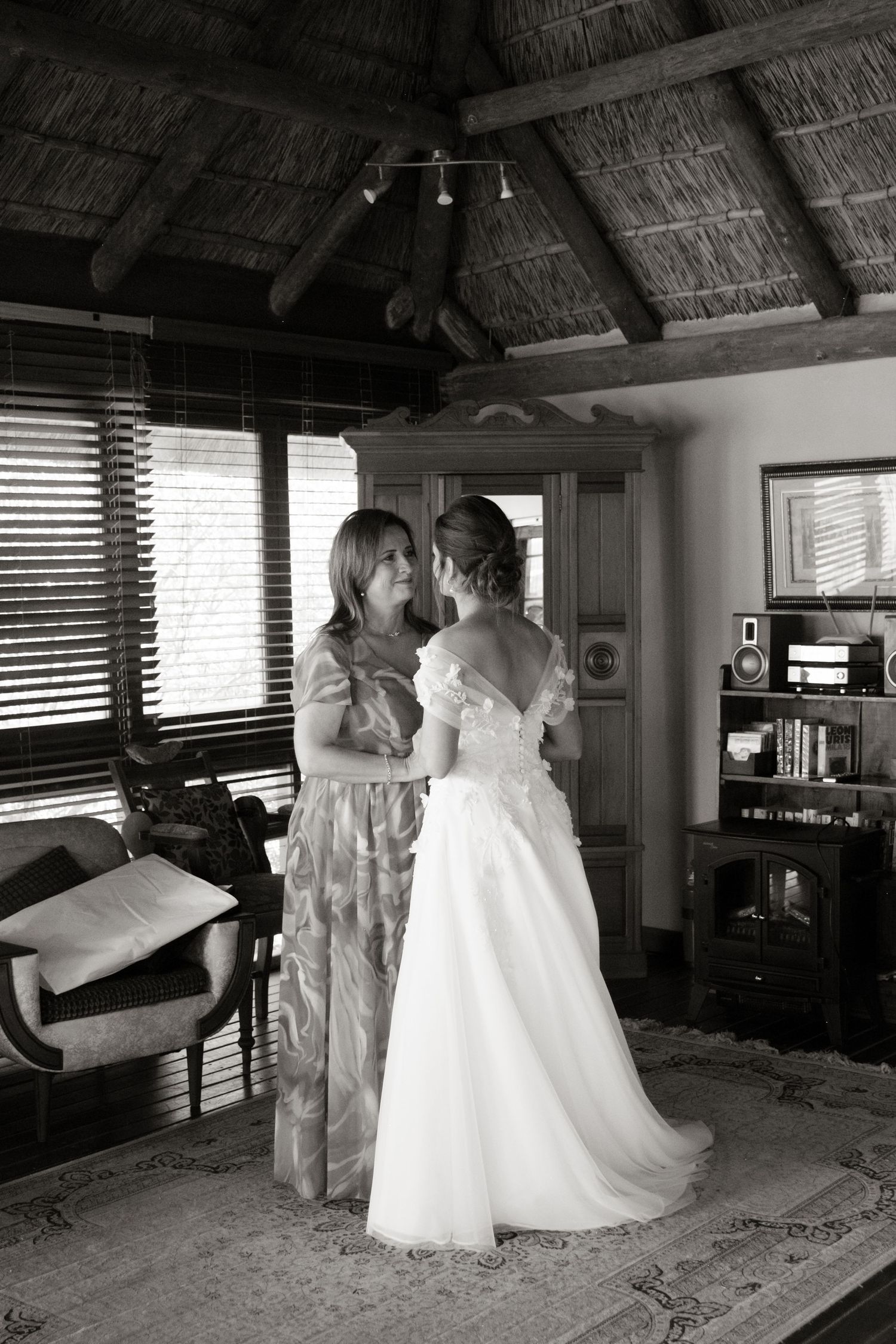Black and white photo of bride in white gown with female guest in rustic thatched roof interior.