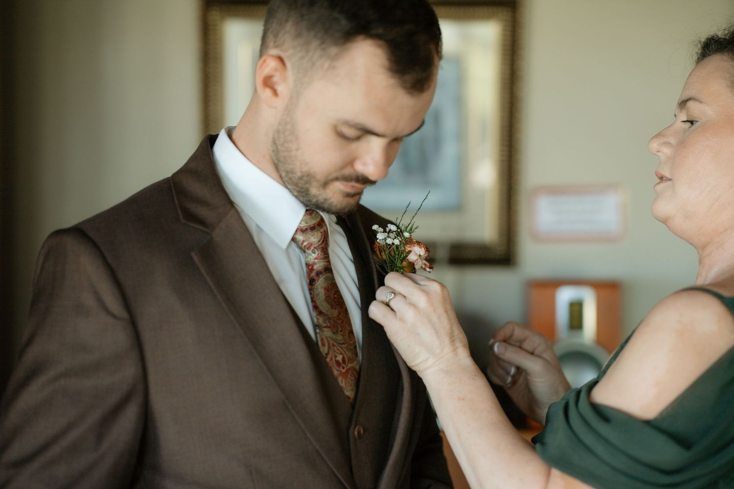 A woman pins a floral boutonniere on a groom in a brown suit and floral tie before his wedding.