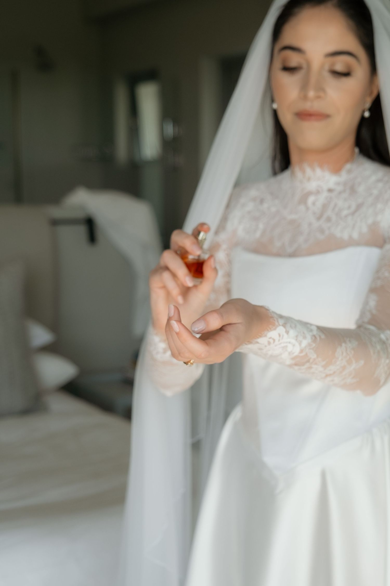 A bride in a white lace wedding gown and veil applies perfume to her wrist while getting ready.